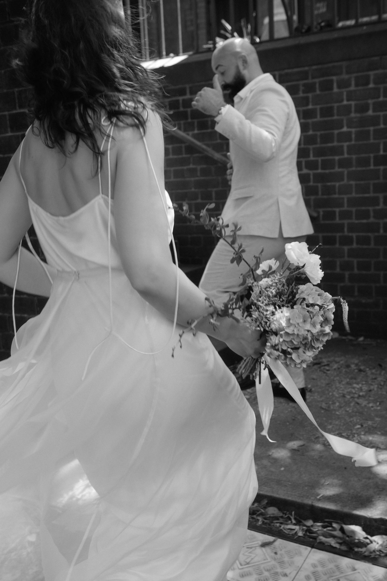 Black and white photo of bride in flowing gown holding bouquet approaching emotional groom in white suit.