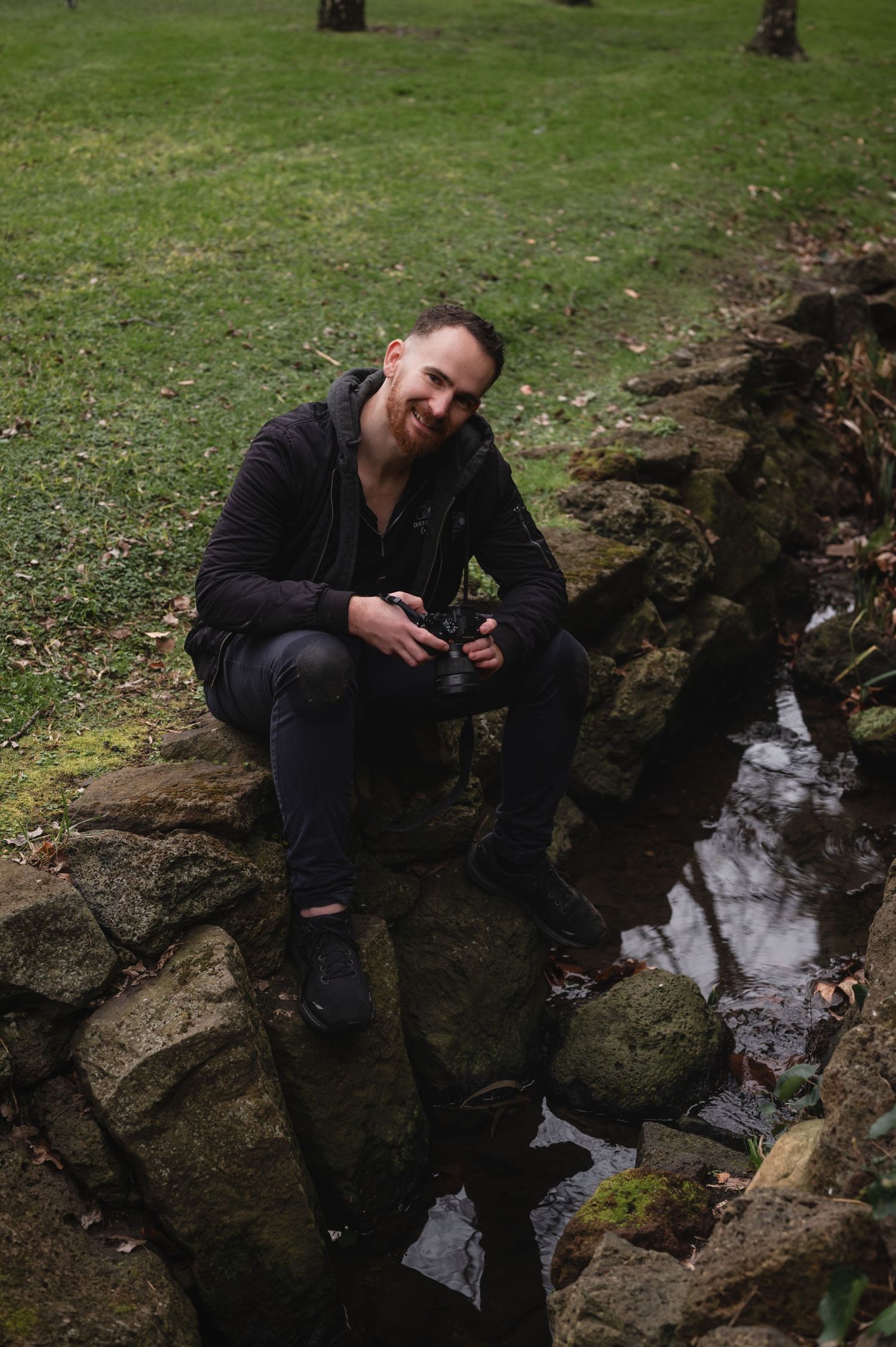 Man in black outfit sitting on mossy rocks by a small stream in a park, holding a camera and smiling.