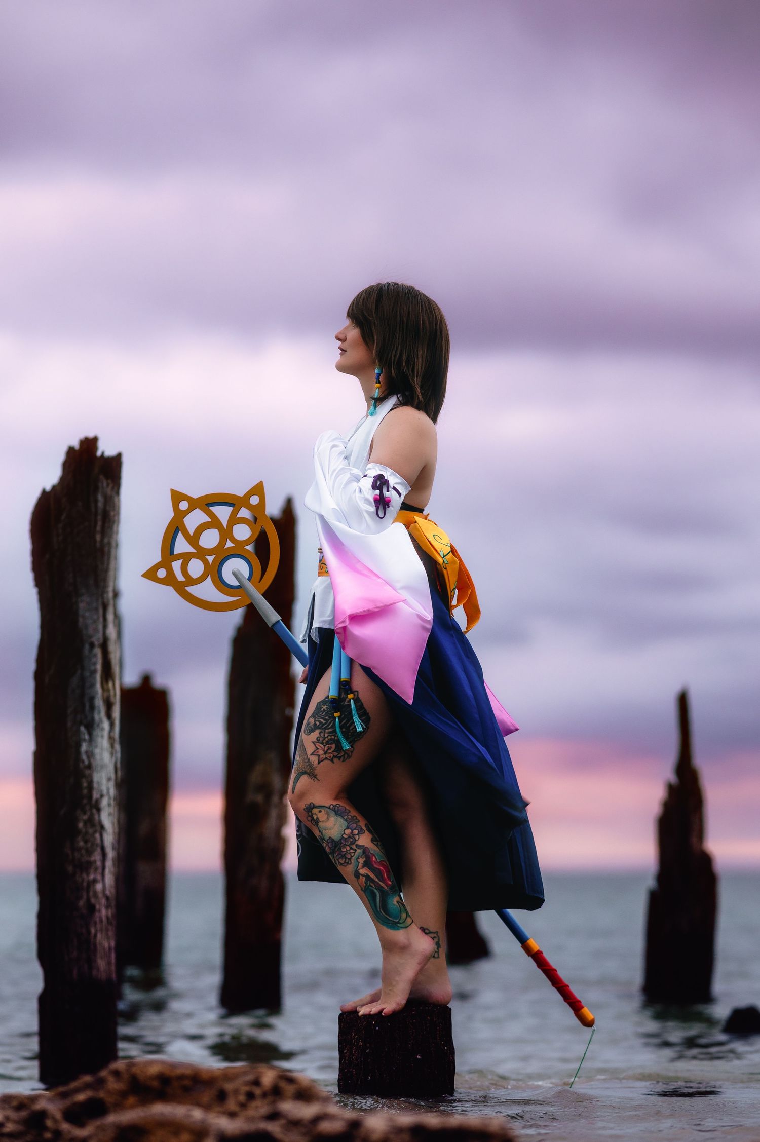 Woman cosplaying as Yuna from Final Fantasy X, standing on wooden posts by the ocean at sunset, holding a staff.