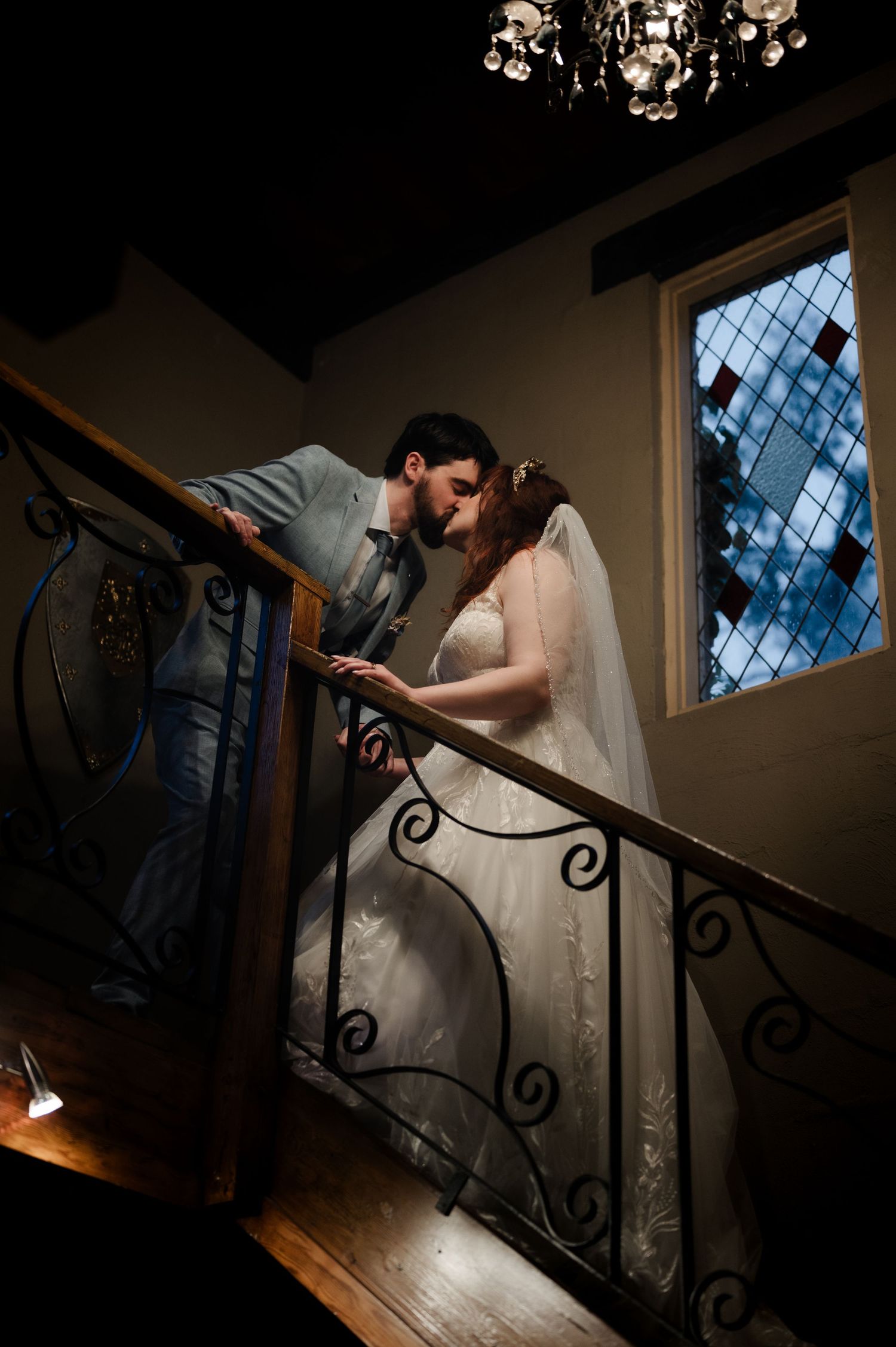 Wedding couple descends ornate wrought iron staircase in dimly lit indoor setting.