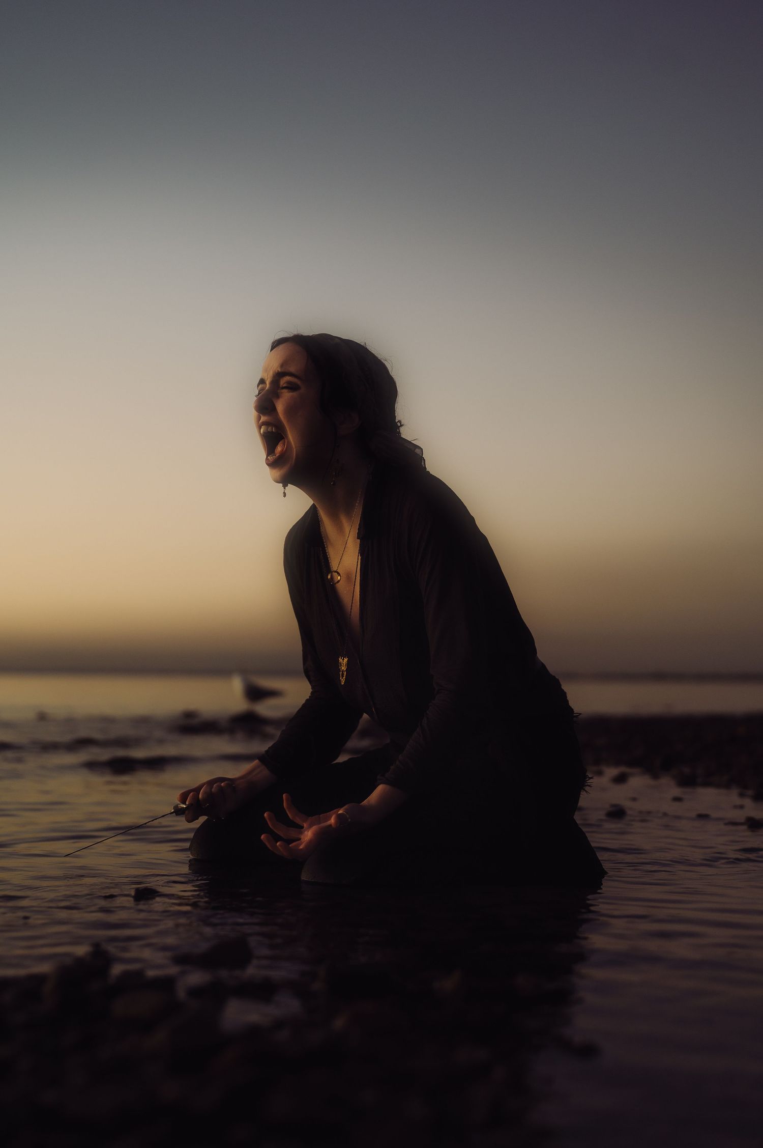 Silhouetted figure sitting by ocean shore at dusk in dramatic lighting.