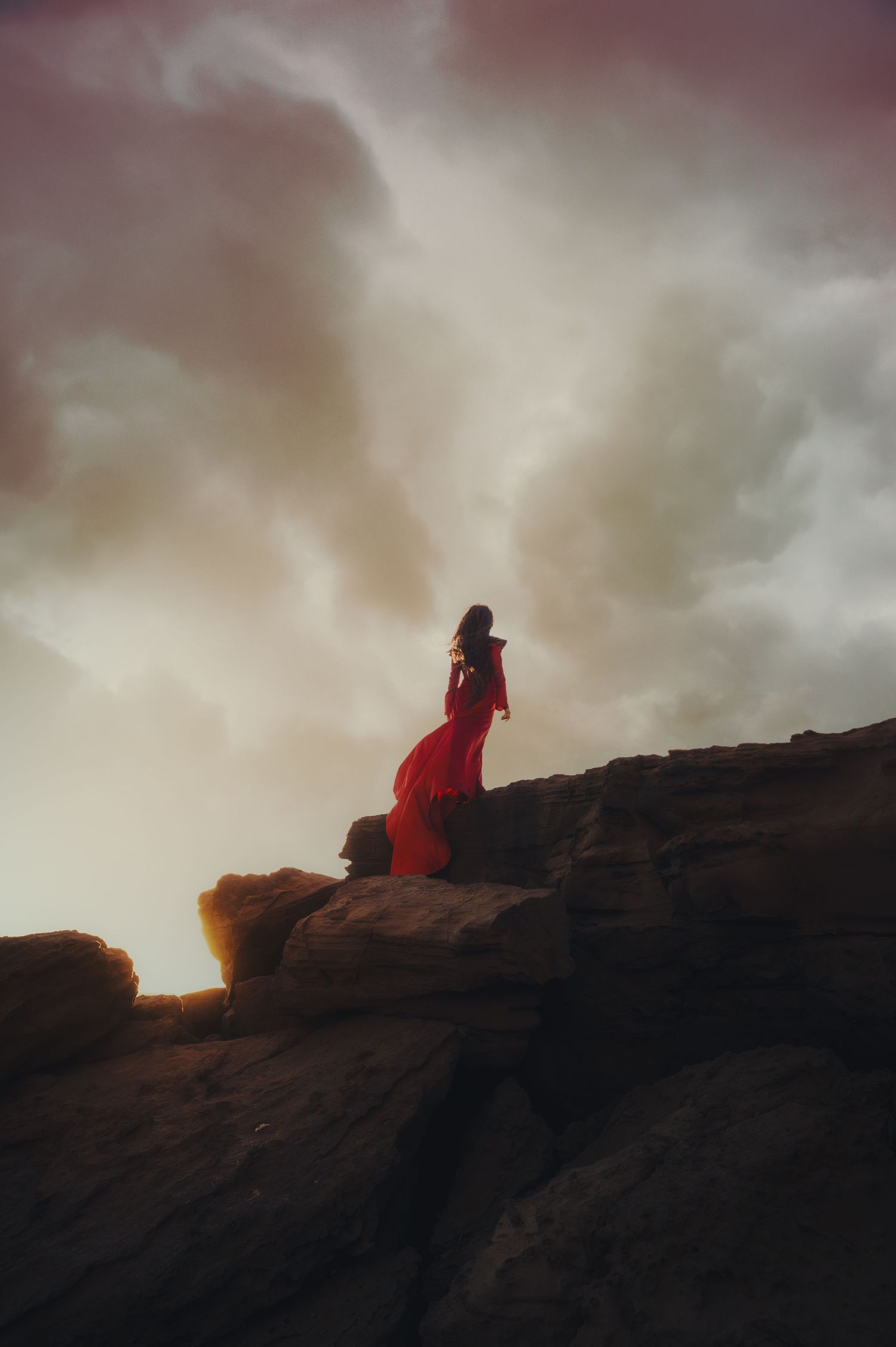 Silhouette in red cloak stands atop rocky cliff at dusk.