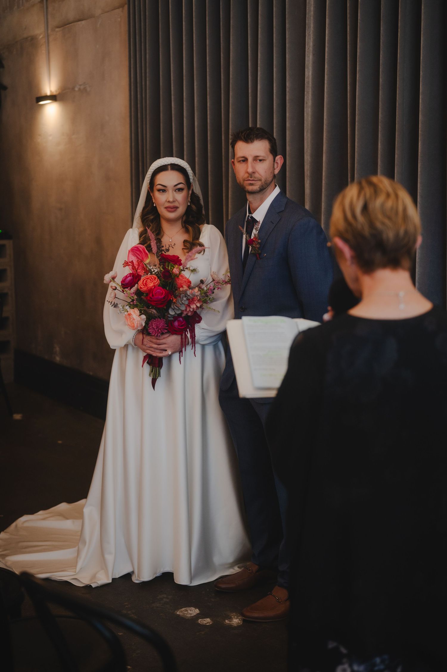 Intimate wedding ceremony in a dimly lit room with a bride in white dress holding red flowers.