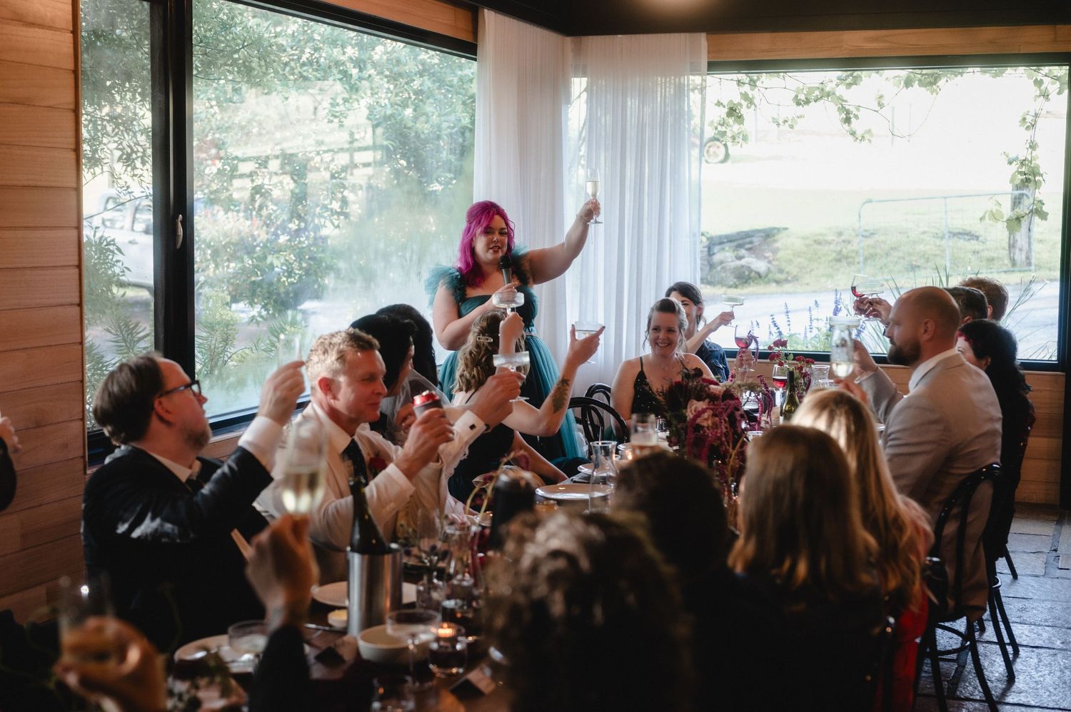 Wedding guests seated in rows at an indoor venue with large windows overlooking a natural outdoor landscape.