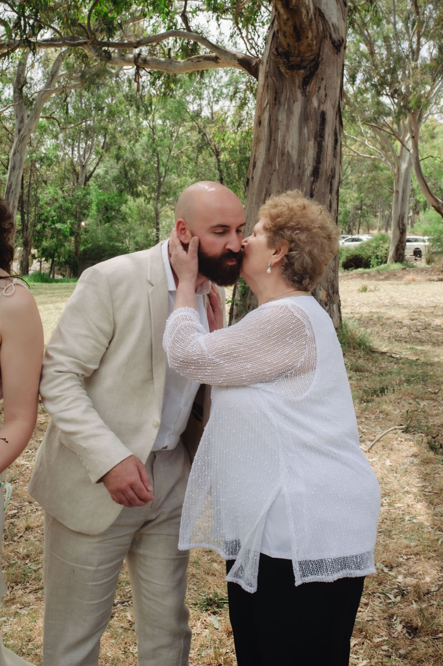 A bald groom in a beige suit shares a tender kiss with an older woman in a white blouse outdoors at a wedding.