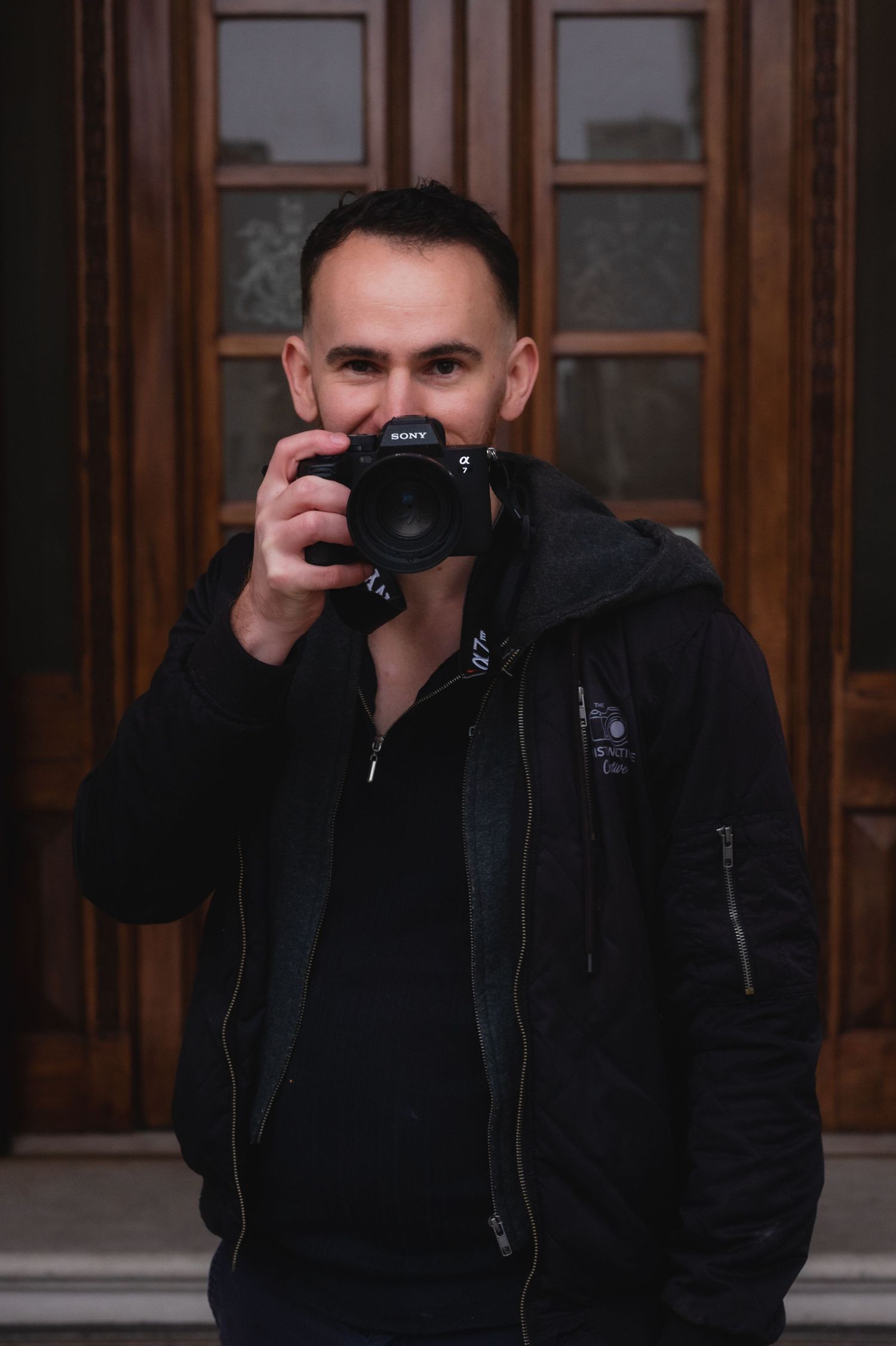 A man in a black jacket holds a Sony camera up to his face while standing in front of wooden doors.