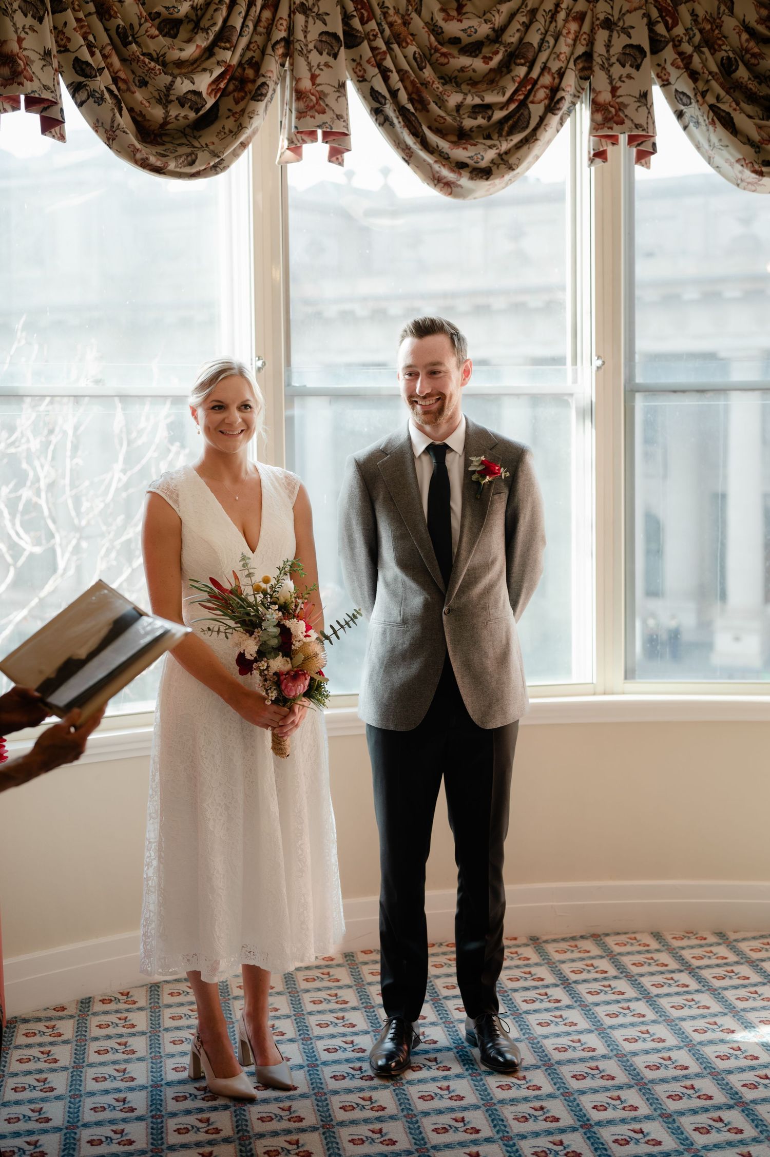 A couple in wedding attire stands by bright windows with elegant curtains and patterned carpet.