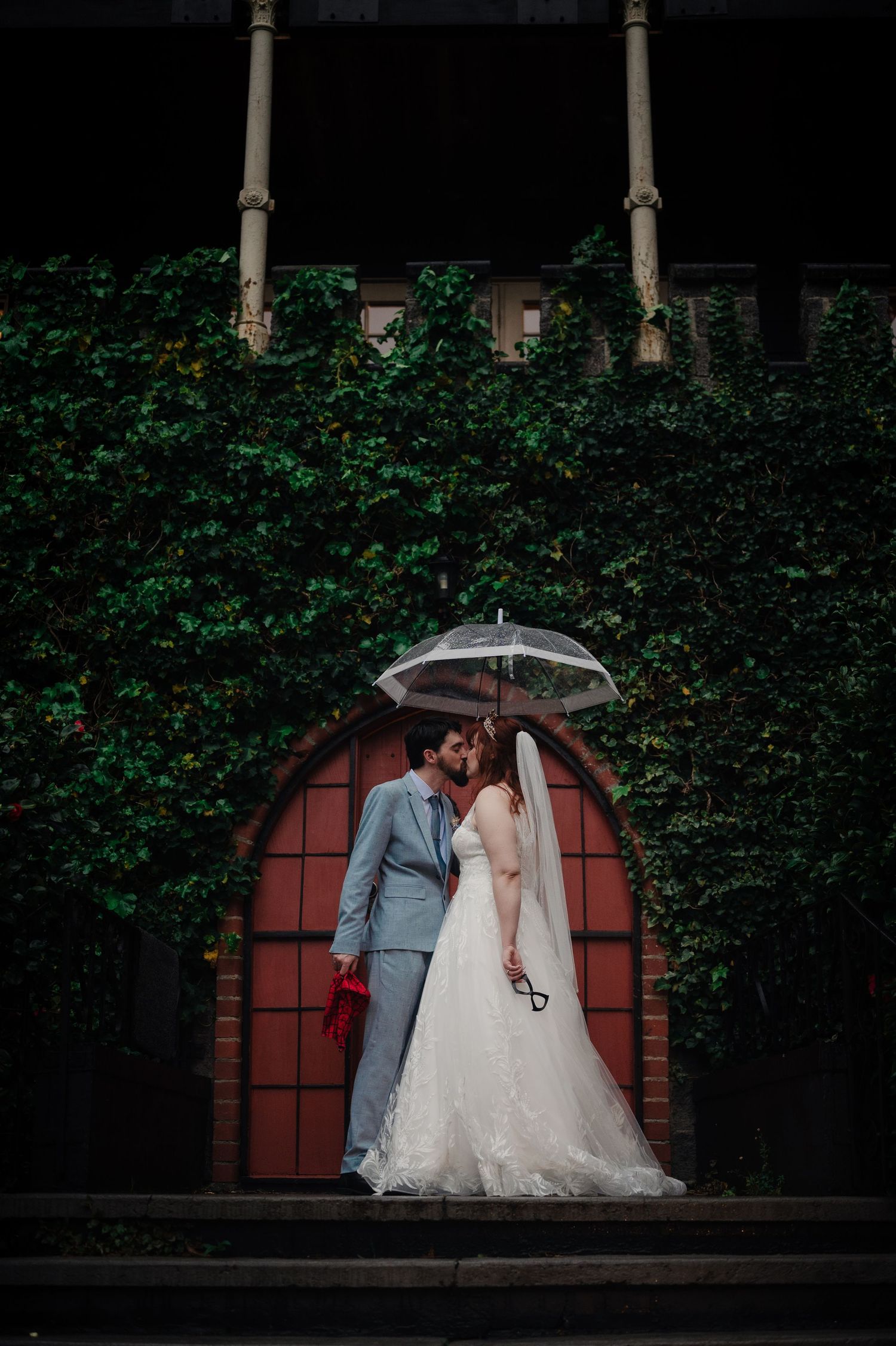 Romantic wedding couple poses together against an ivy-covered wall with red archway in garden setting.