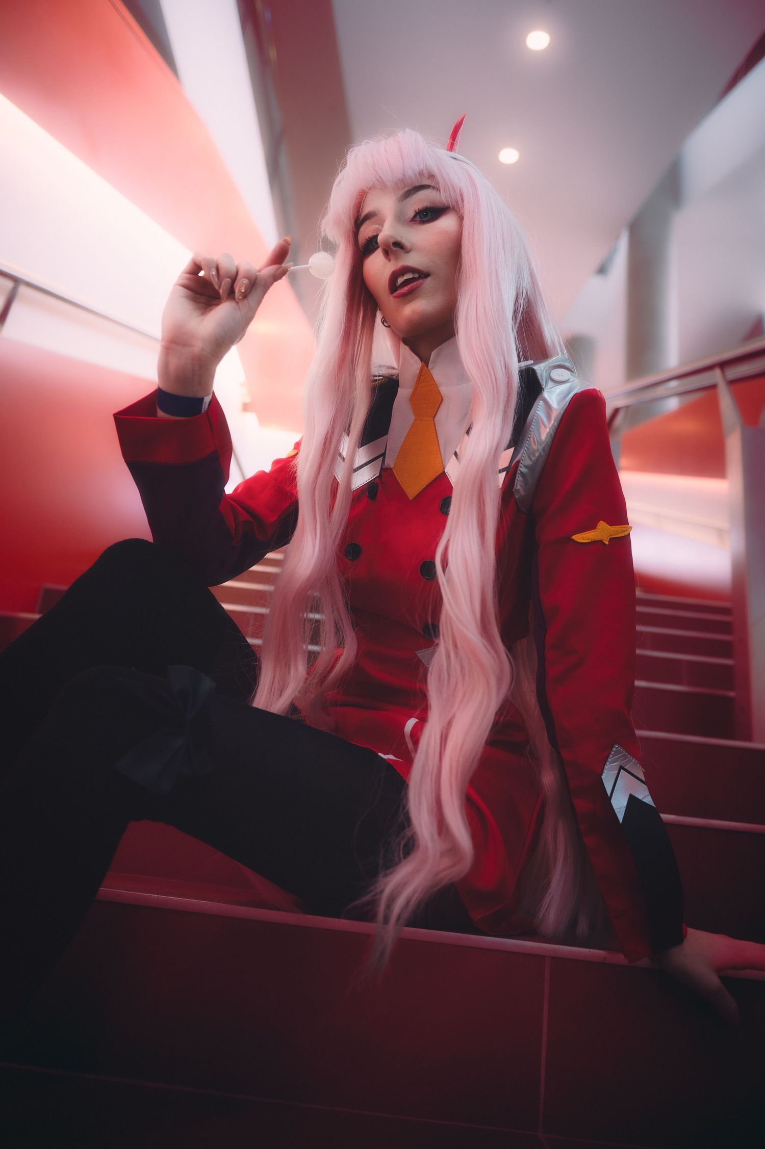 Cosplay photography in red-lit interior with long pink hair and dramatic poses.