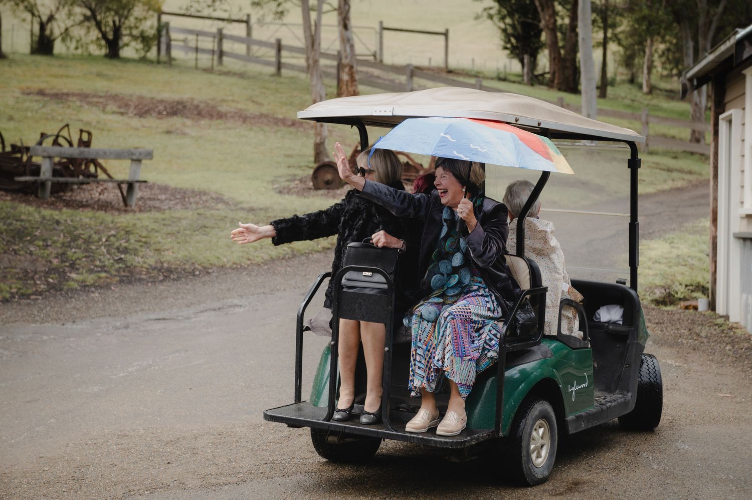 Group of women riding a green golf cart in the rain, one holding a colorful umbrella, pointing ahead on a rural path.