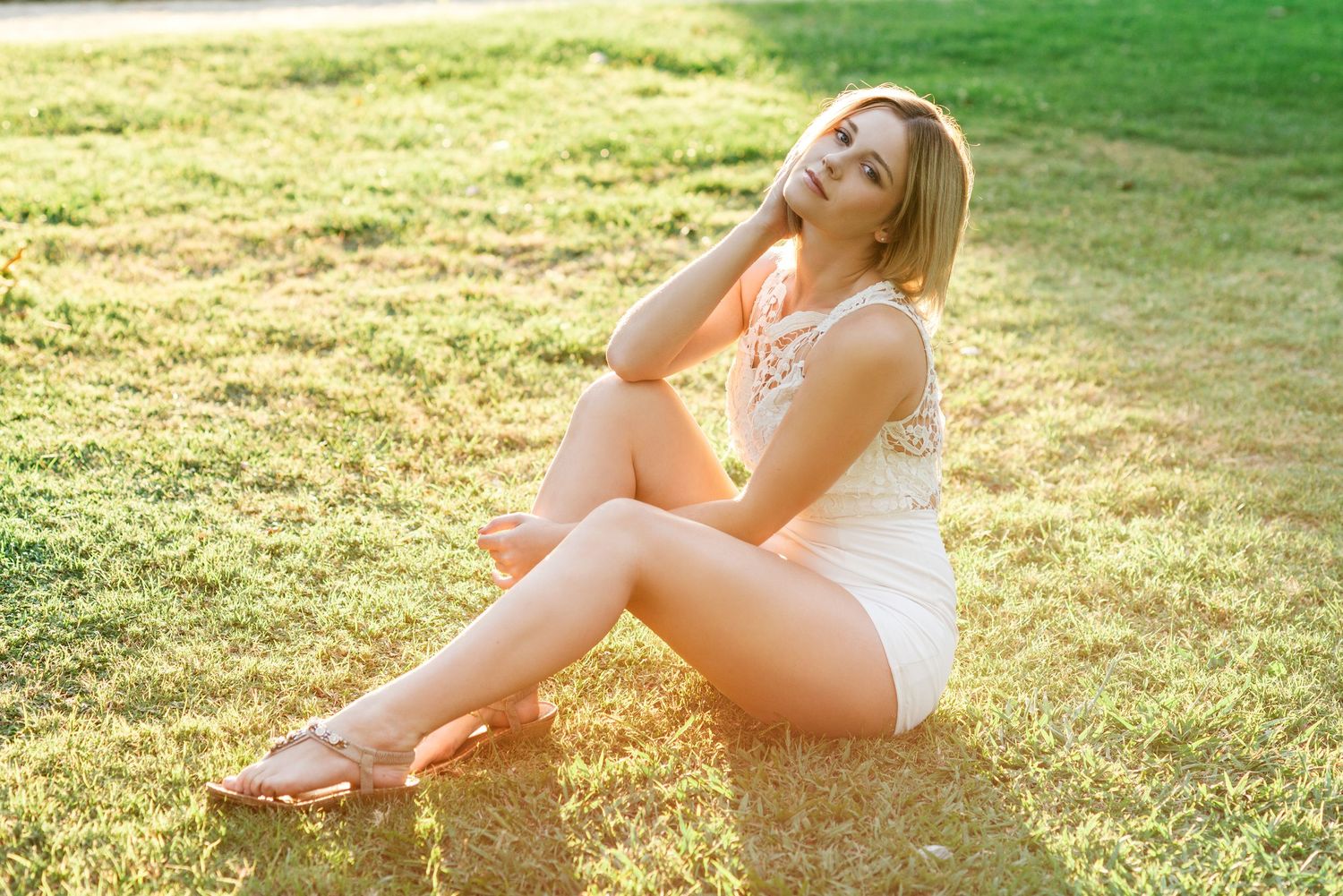 Young blonde woman in white romper sitting on green grass in warm sunlight, posing with hand on neck.