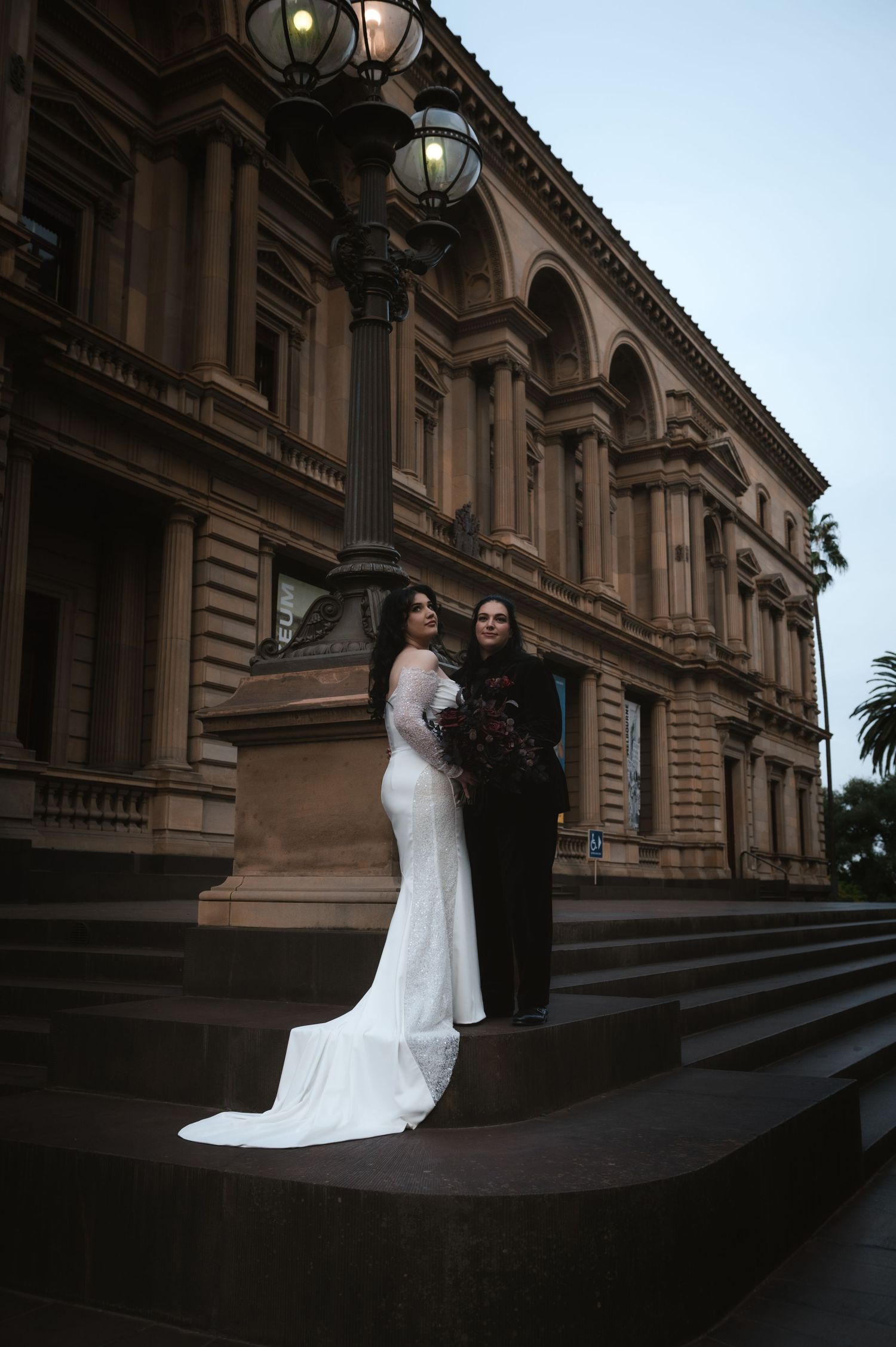 A wedding couple poses on grand stone steps of a historic building during sunset.