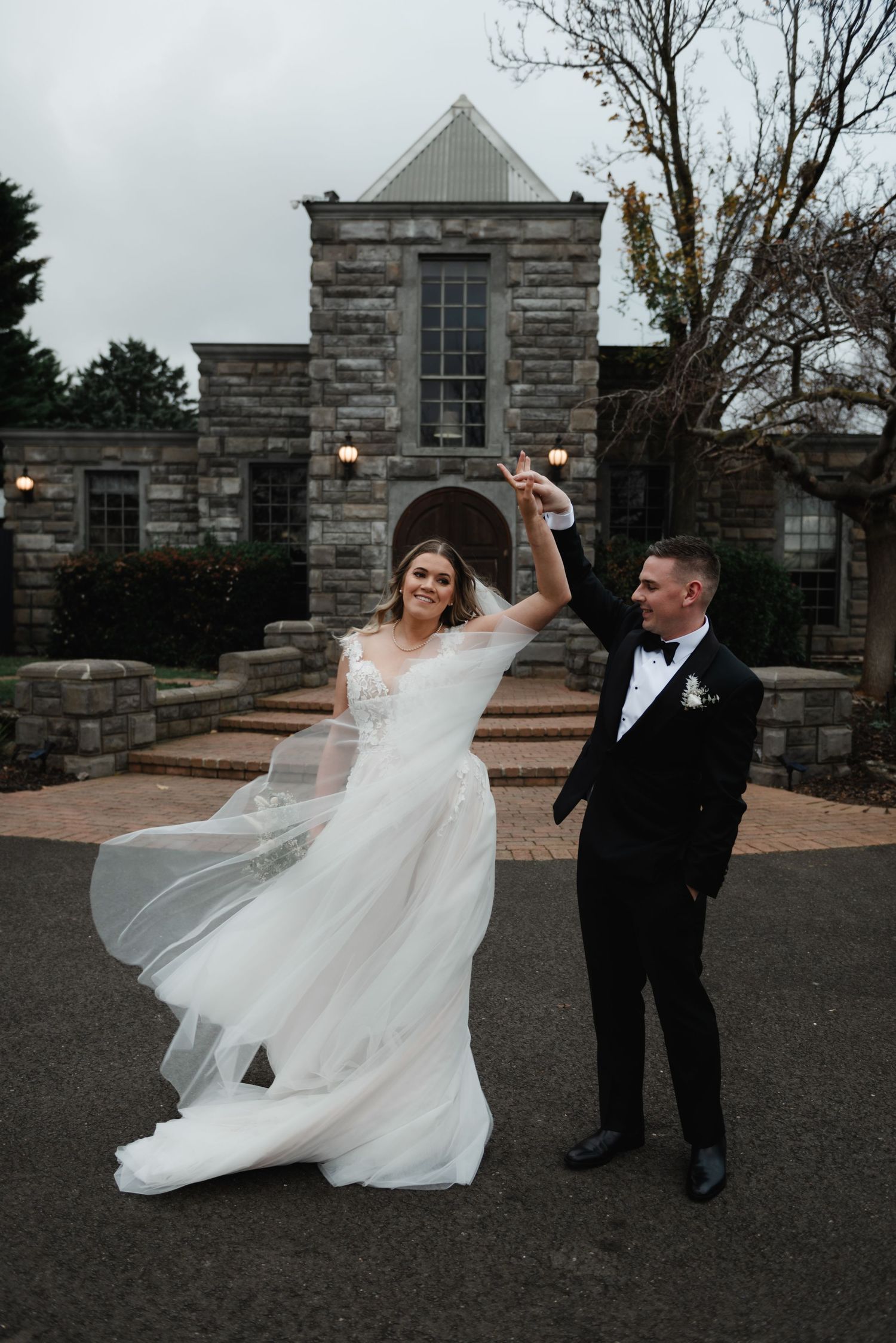Wedding couple dances playfully with flowing dress in front of stone building.