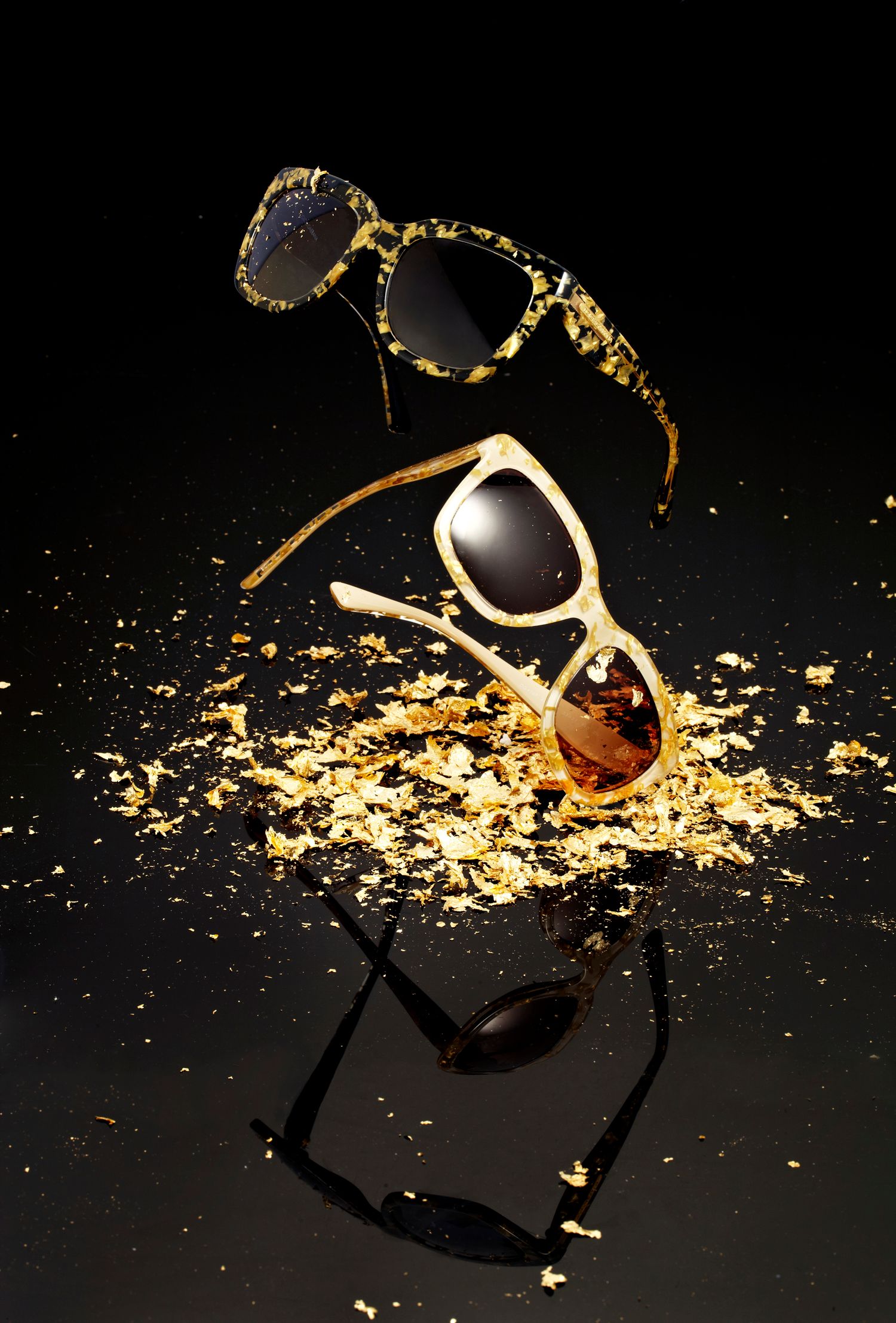 Two luxury sunglasses with gold flake embellishments displayed dramatically on a reflective black surface.
