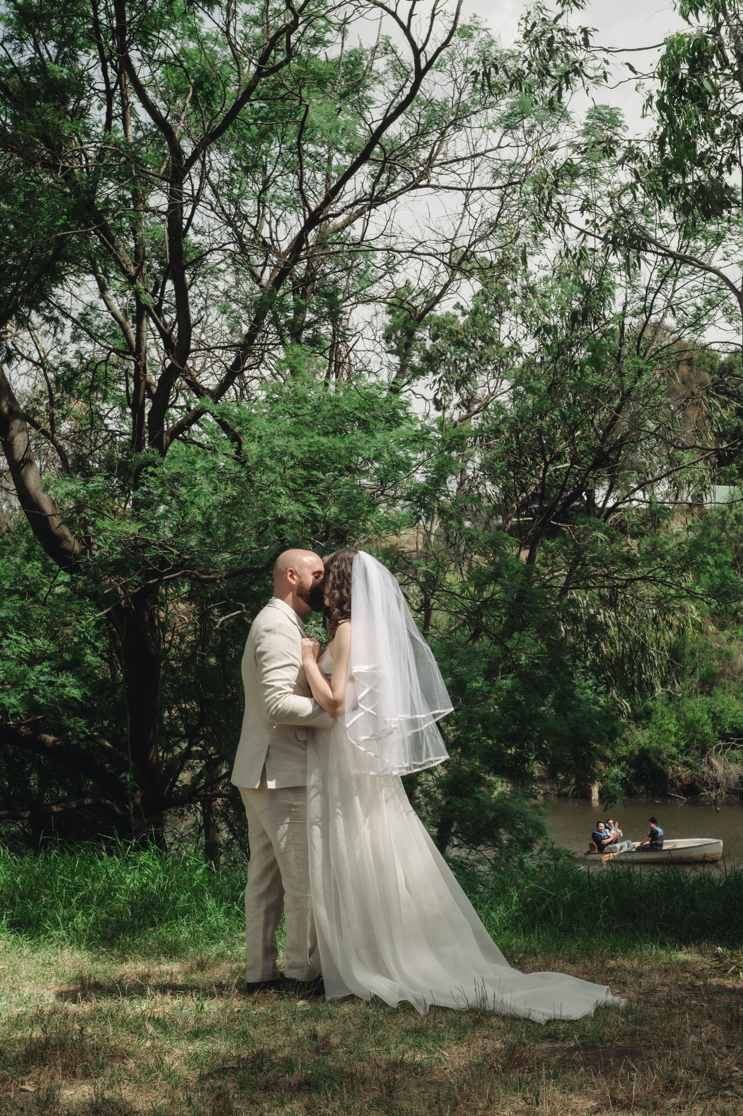 Bride and groom share a romantic kiss outdoors near a river, surrounded by lush green trees on their wedding day.