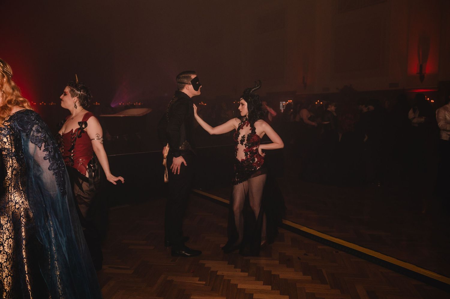 Two masked guests dance at a dimly lit masquerade ball in elegant costumes.