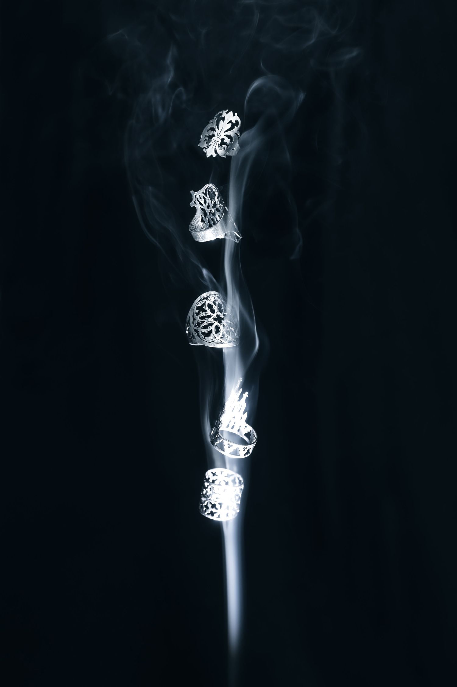 Artistic smoke photography of multiple ornate rings creating a vertical line against black background.