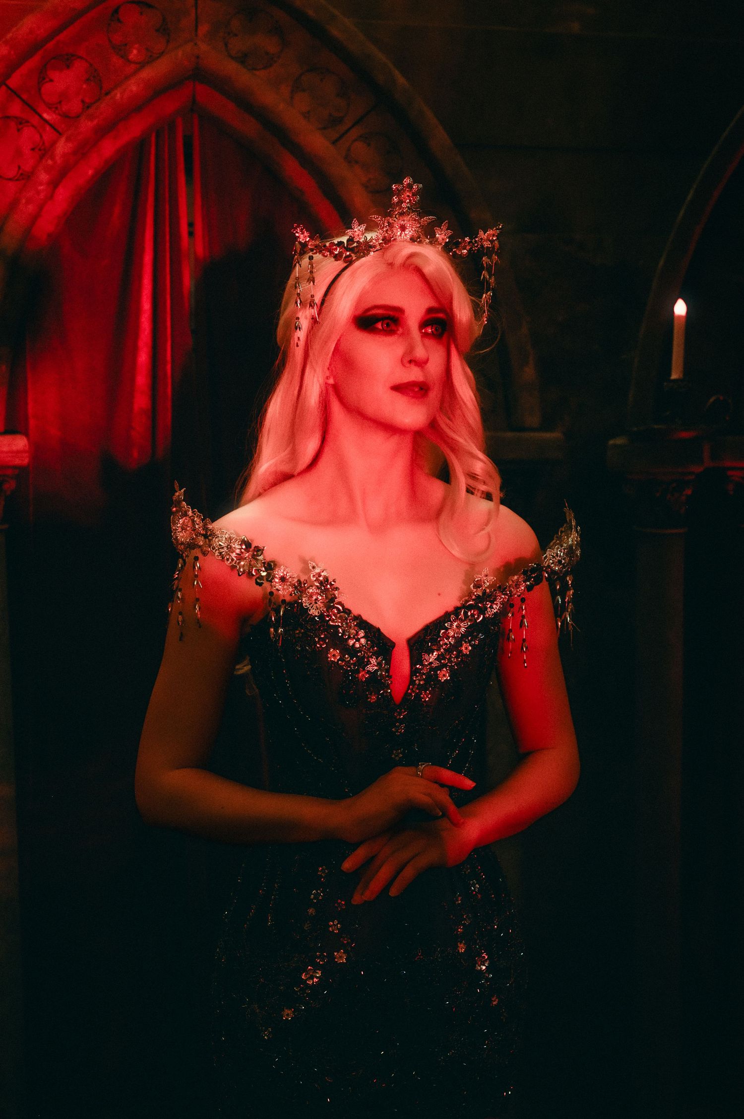 A blonde woman in a dark floral gown and ornate crown poses dramatically in a dimly lit gothic castle setting.