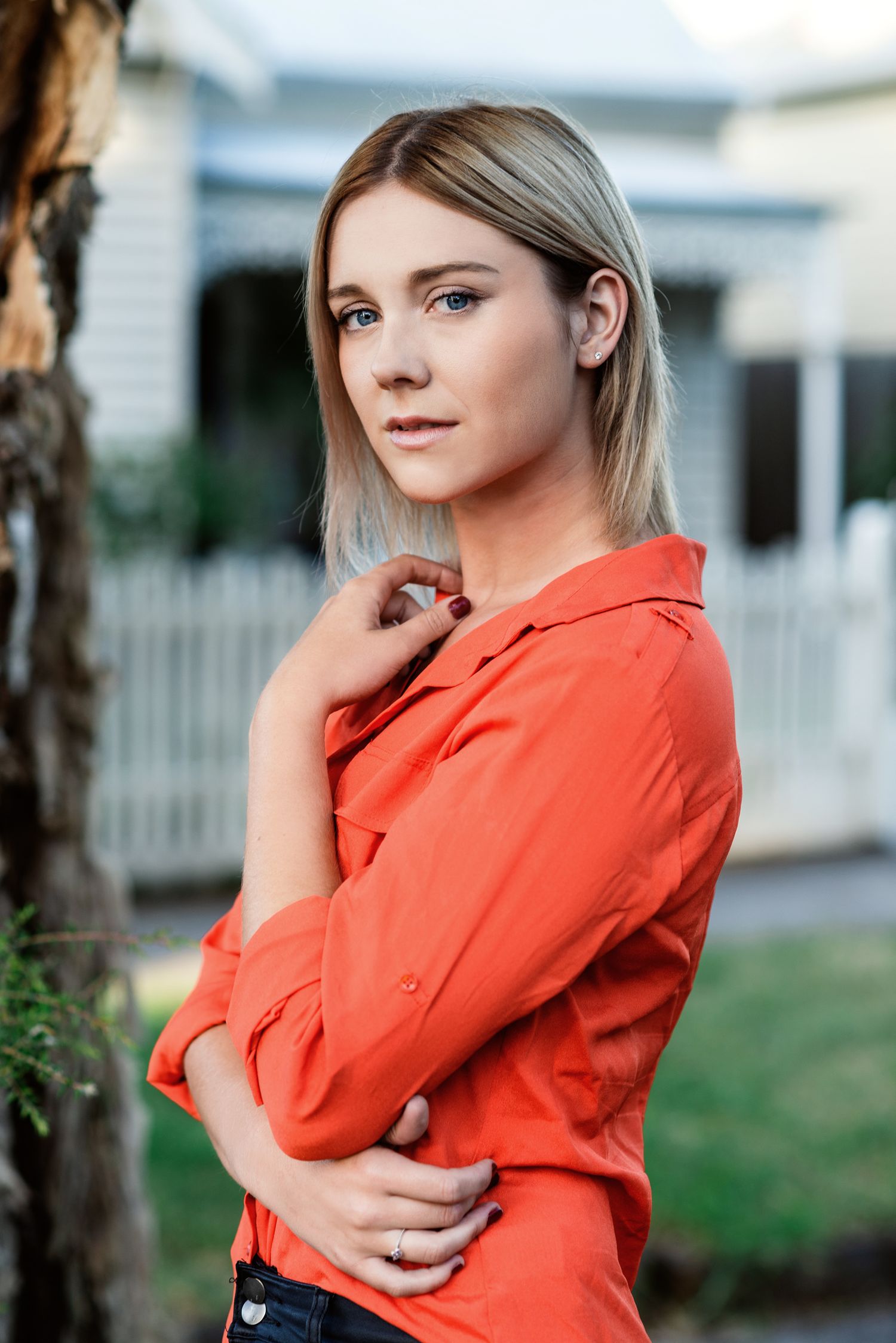 Young blonde woman in orange shirt poses outdoors near white picket fence, looking thoughtfully at camera.