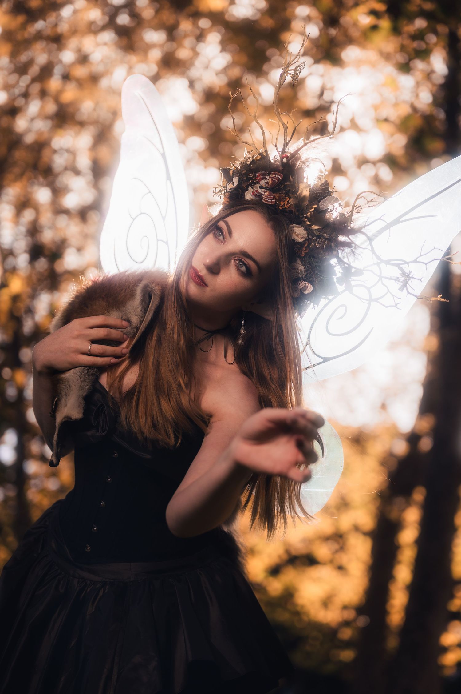 Woman in black corset dress with fairy wings and floral crown reaches toward camera in autumn forest.