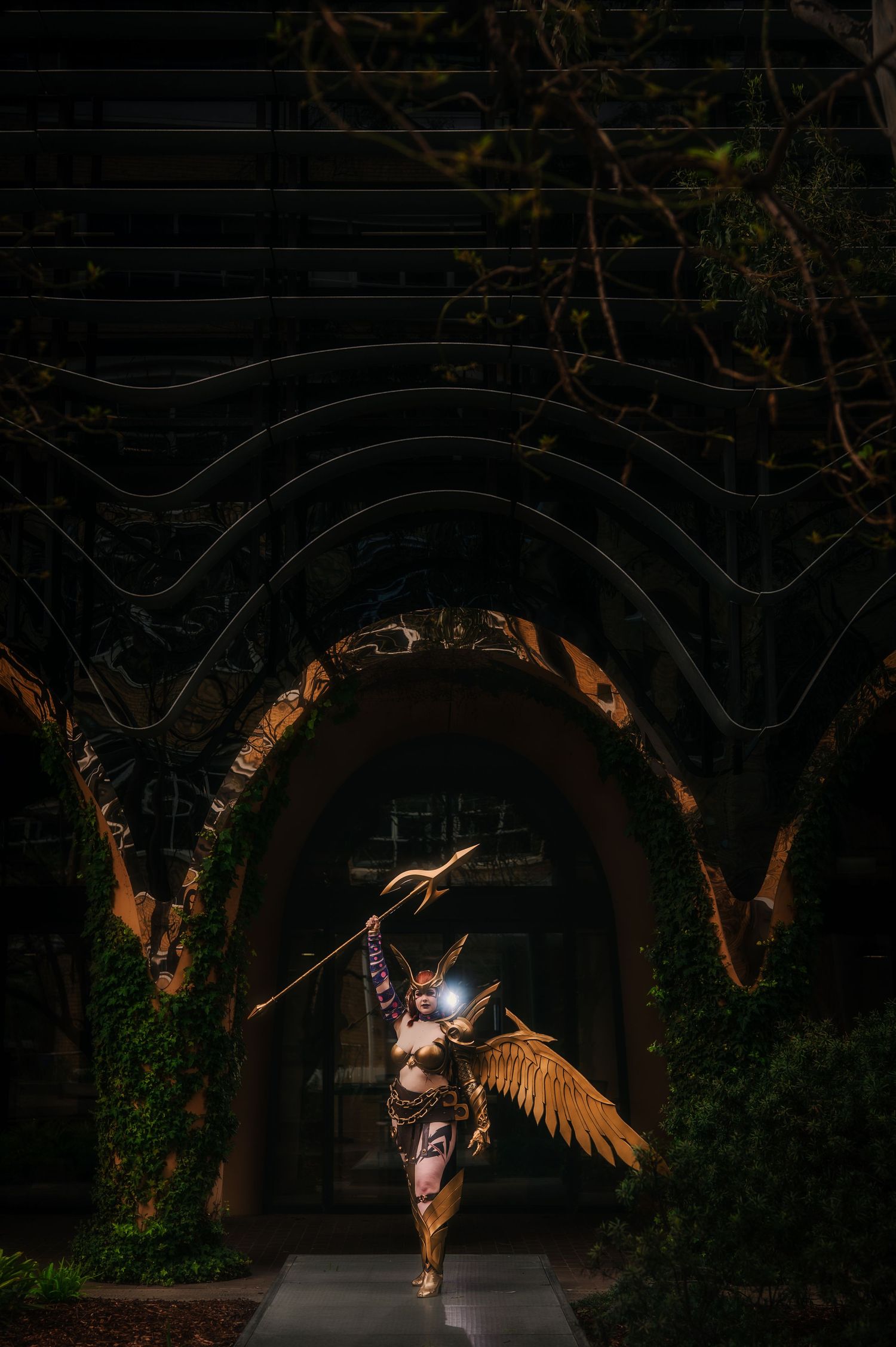 A cosplayer dressed as a golden armored angel with wings and spear poses under an arched entrance at night.