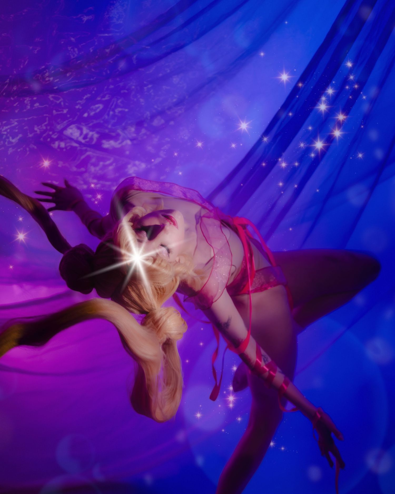 A performer in red lingerie poses dramatically under blue sparkly draping fabric with magical star light effects.