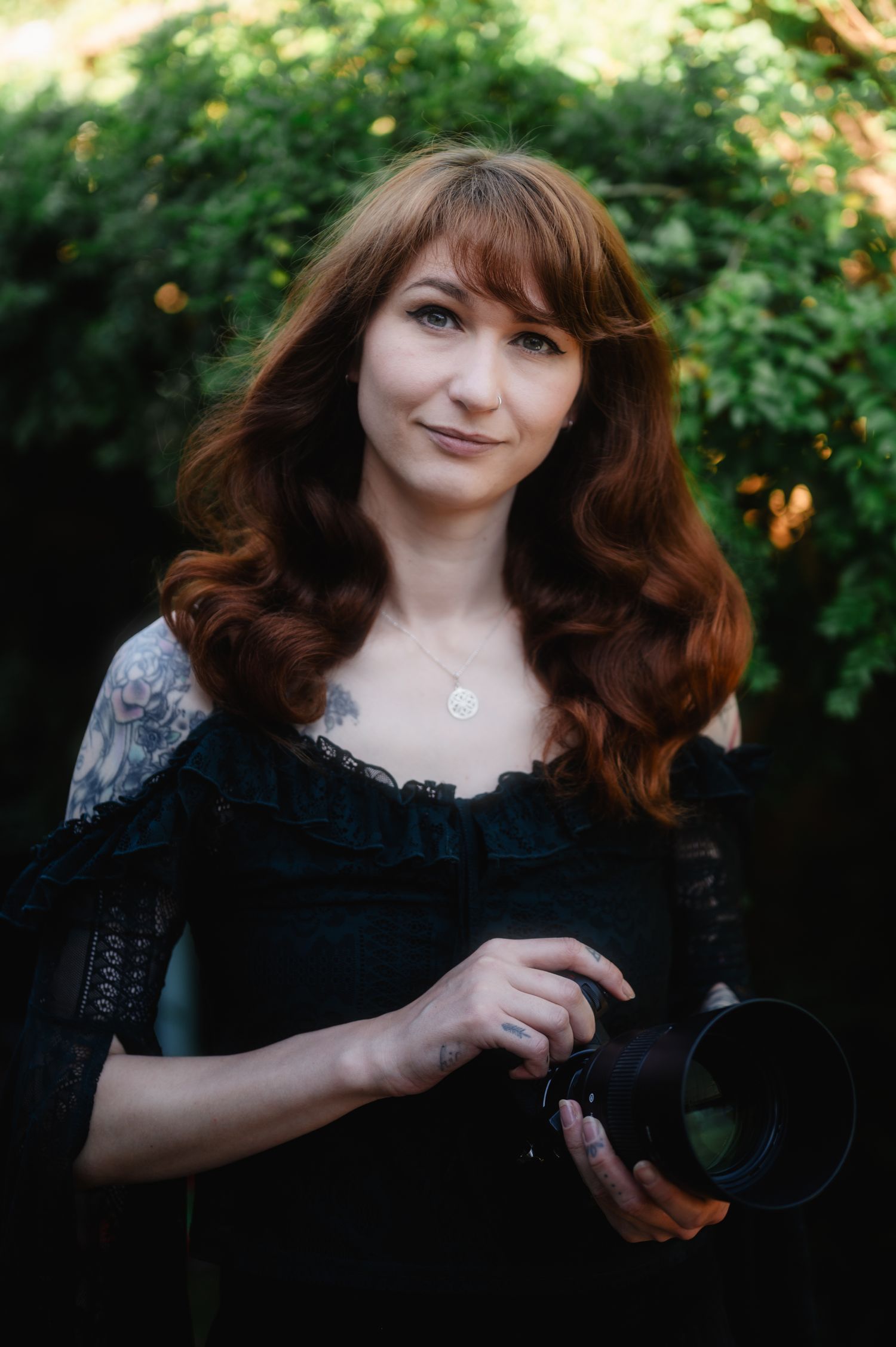 Portrait of a photographer in a black lace dress holding a camera outdoors with green foliage in the background.