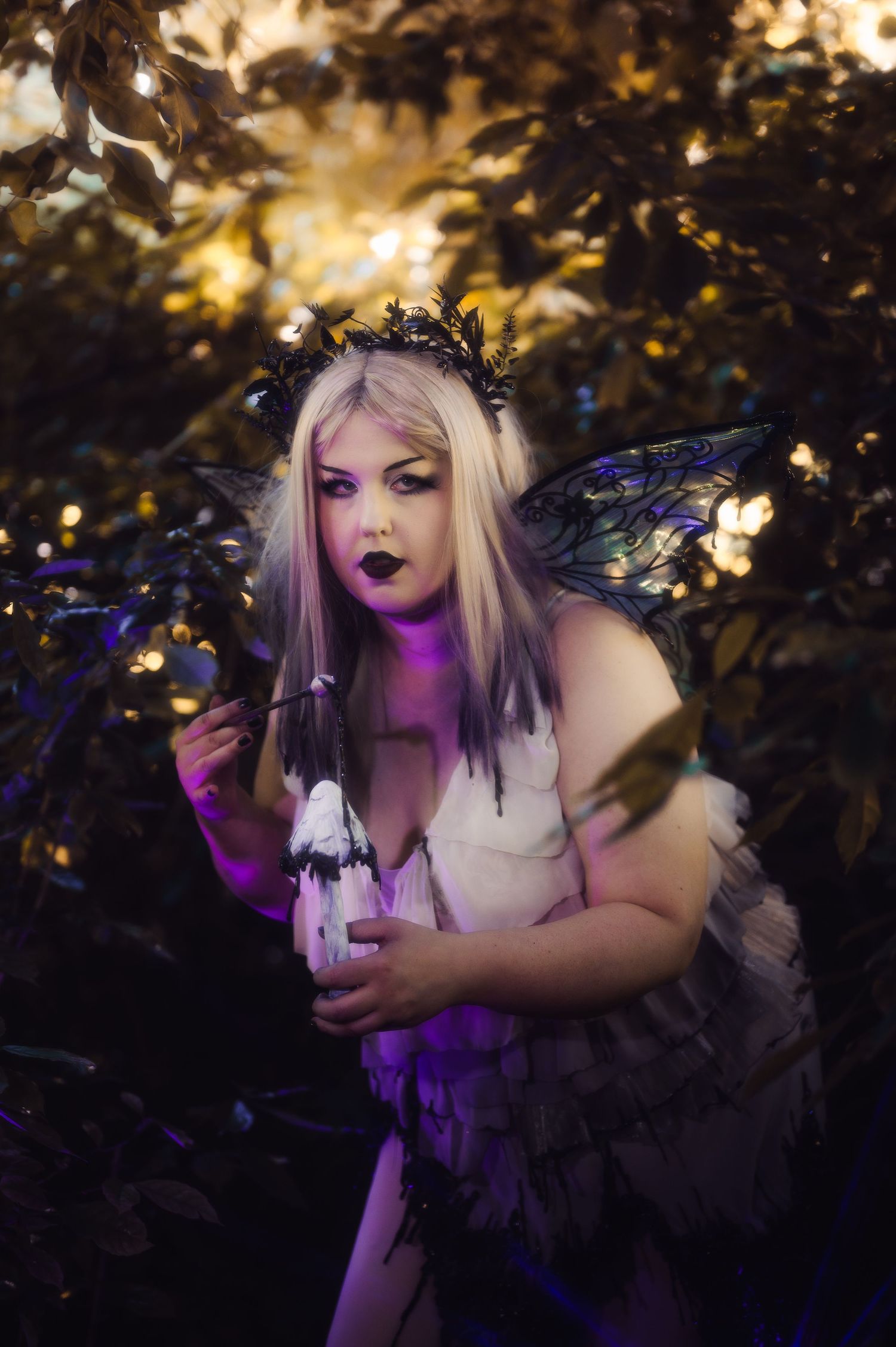 A woman dressed as a dark fairy with black wings, floral crown, and gothic makeup poses among glowing forest foliage.