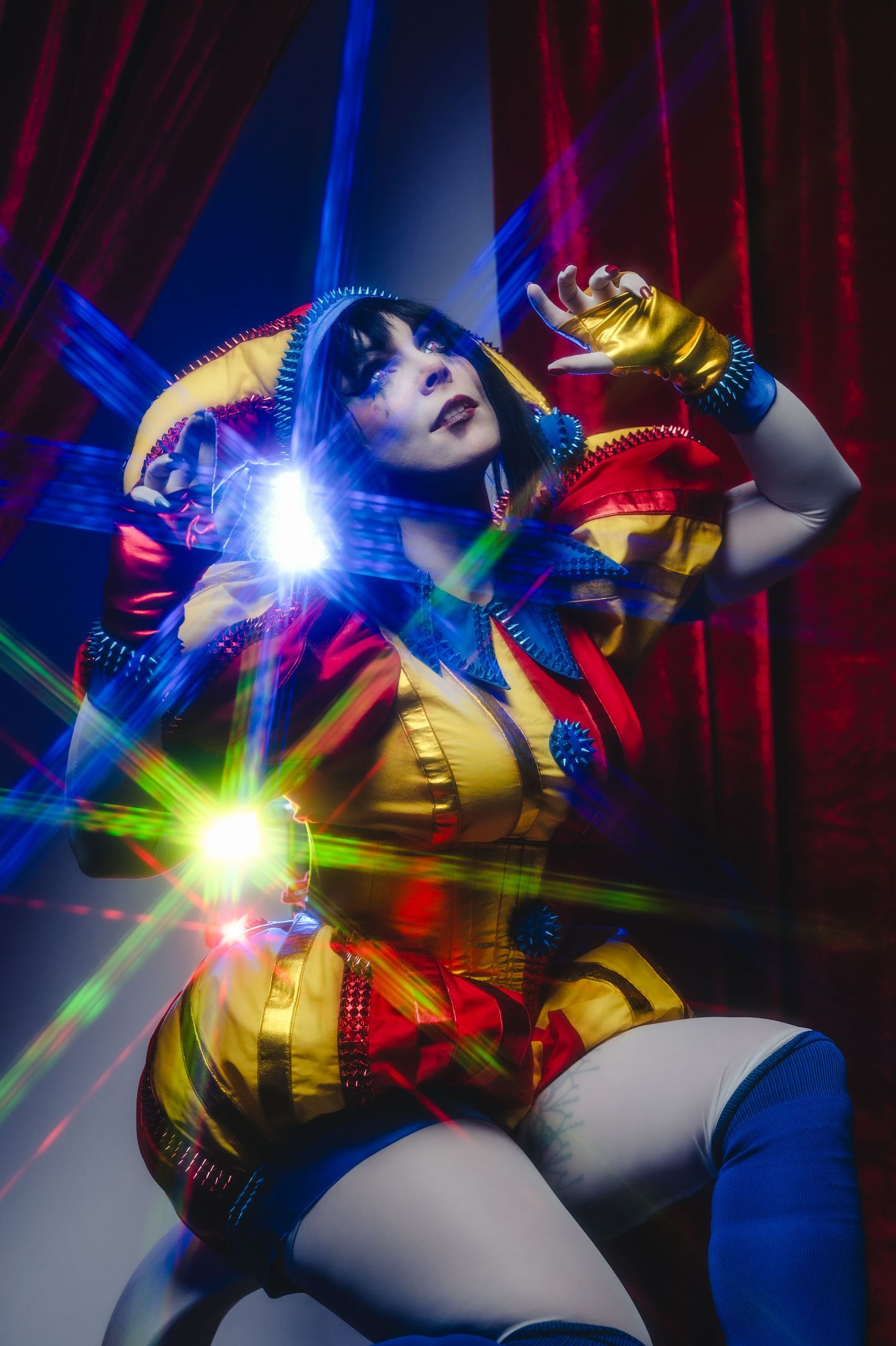 Colorful cosplay performer in gold and red costume with dramatic laser light effects against red curtain backdrop.