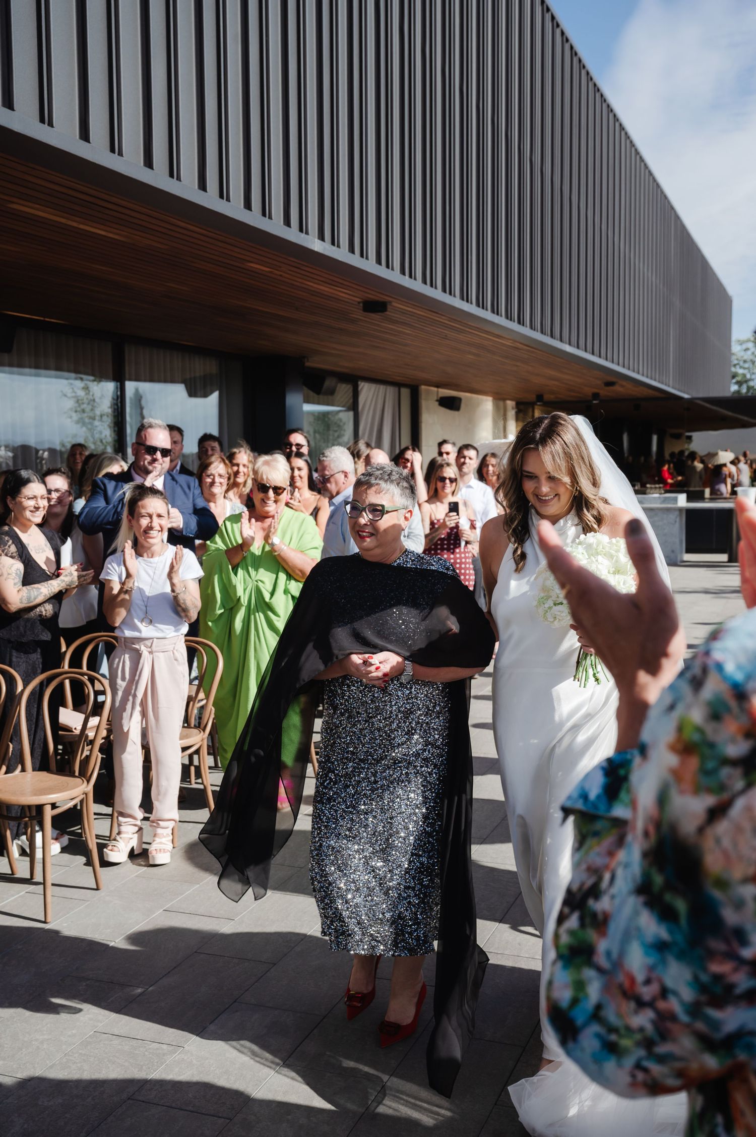Wedding guests and celebrants gathered outside a modern building with vertical architectural details.