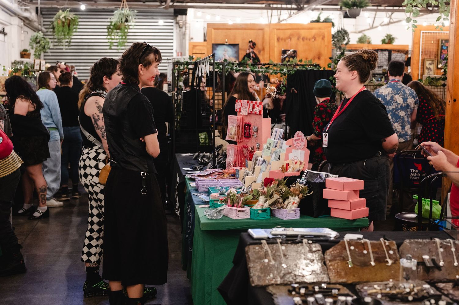 A busy indoor craft market or vendor fair with multiple tables displaying handmade goods and shoppers browsing merchandise.