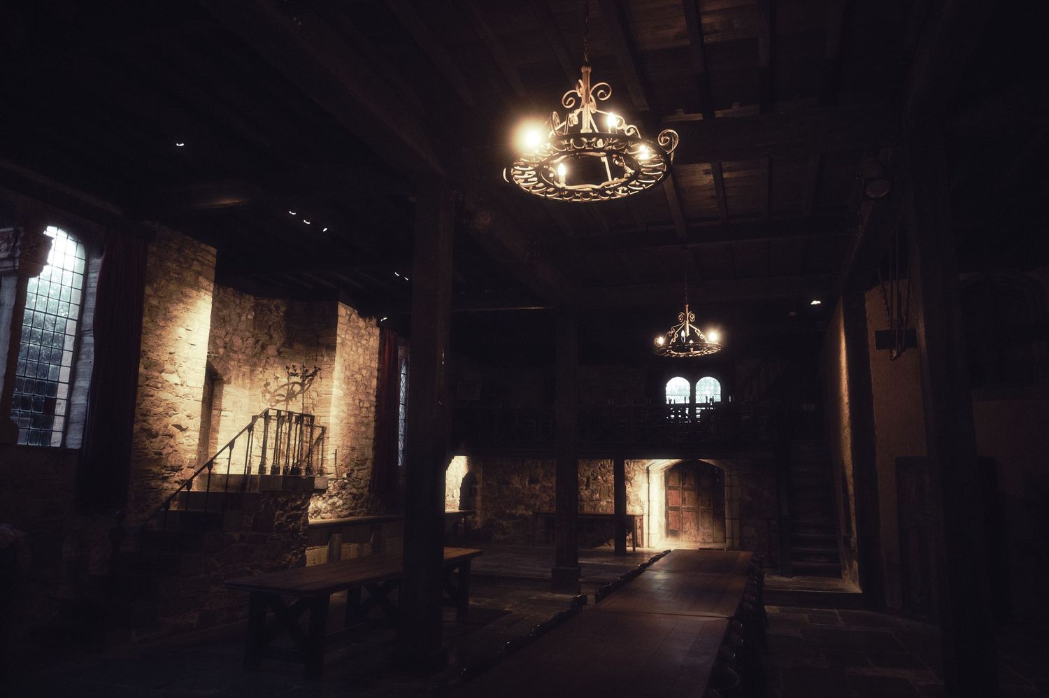 Dark medieval stone hall interior with iron chandeliers and arched windows casting dim light.