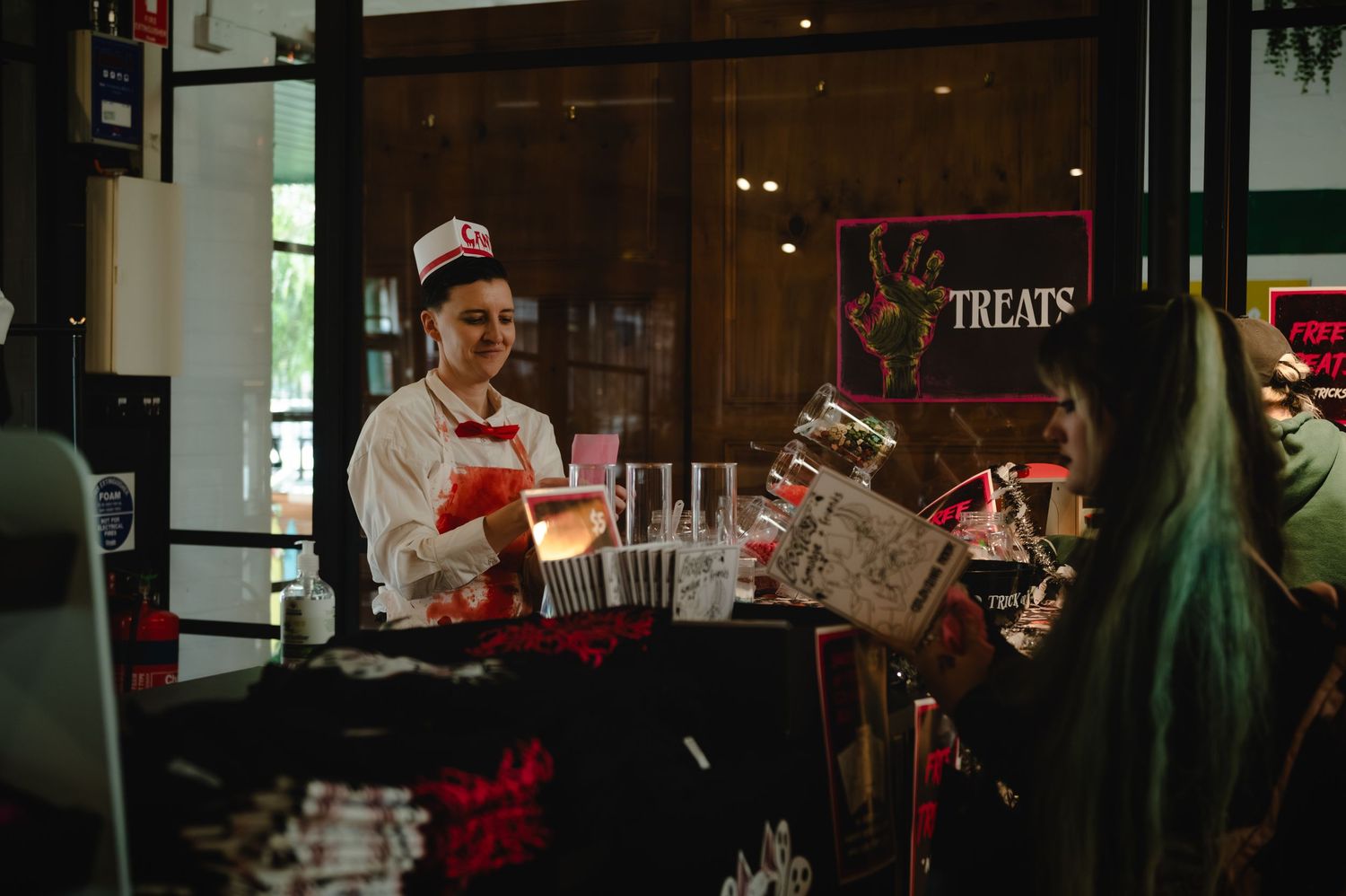 A smiling server in retro uniform serves drinks at a treats booth during a festive indoor event.