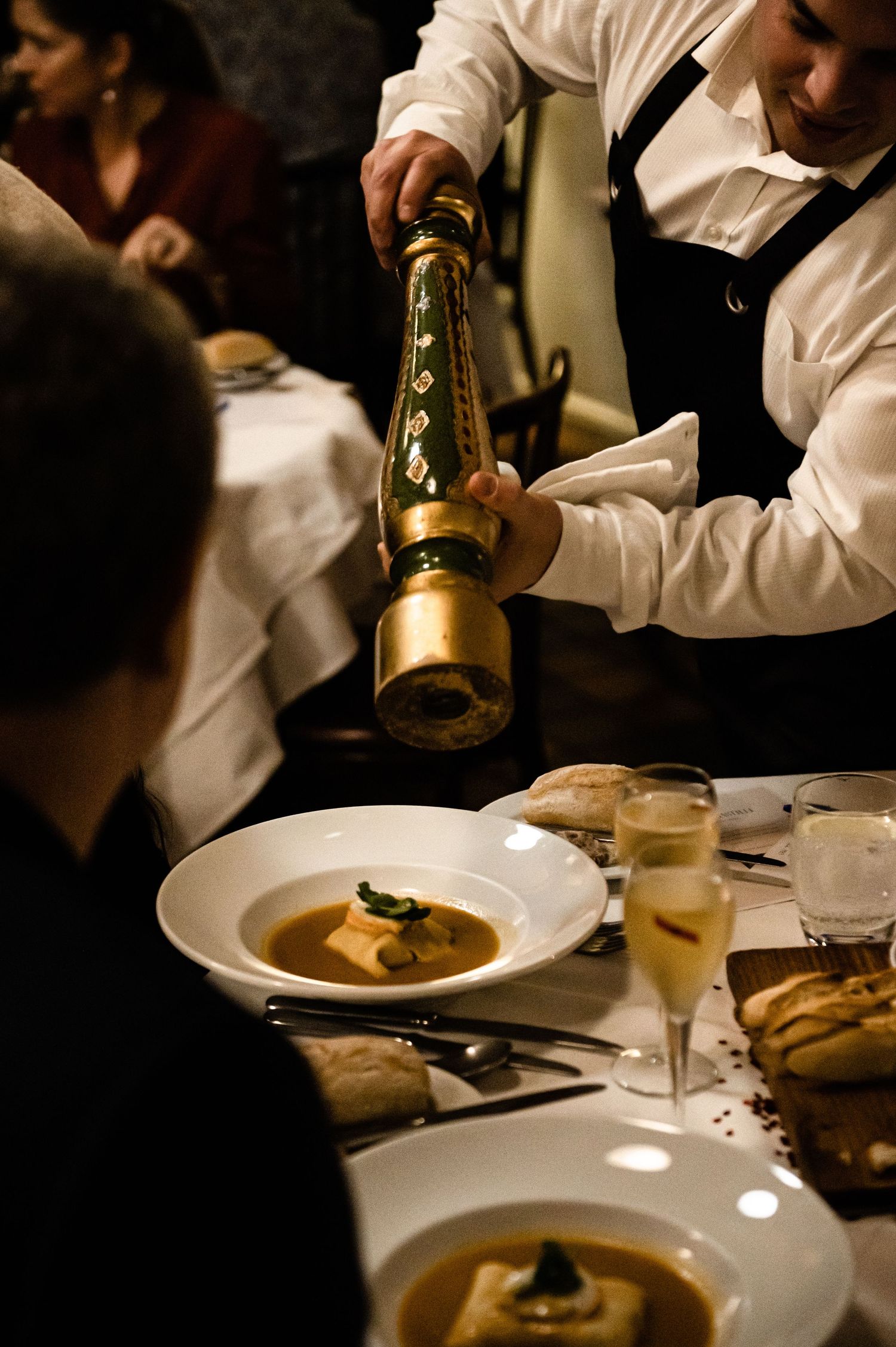 Waiter using an ornate gold pepper grinder to season soup at an upscale restaurant dinner table.