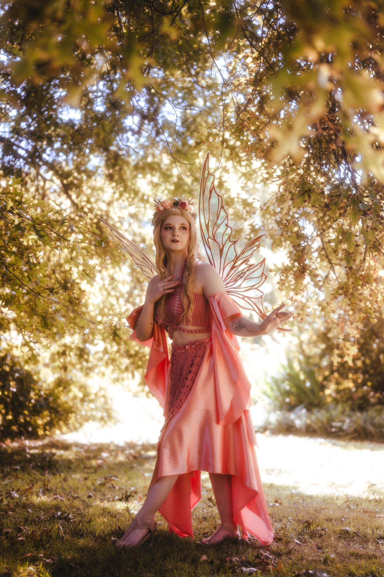 A woman in a pink fairy costume with wings and floral crown poses in a sunlit forest.