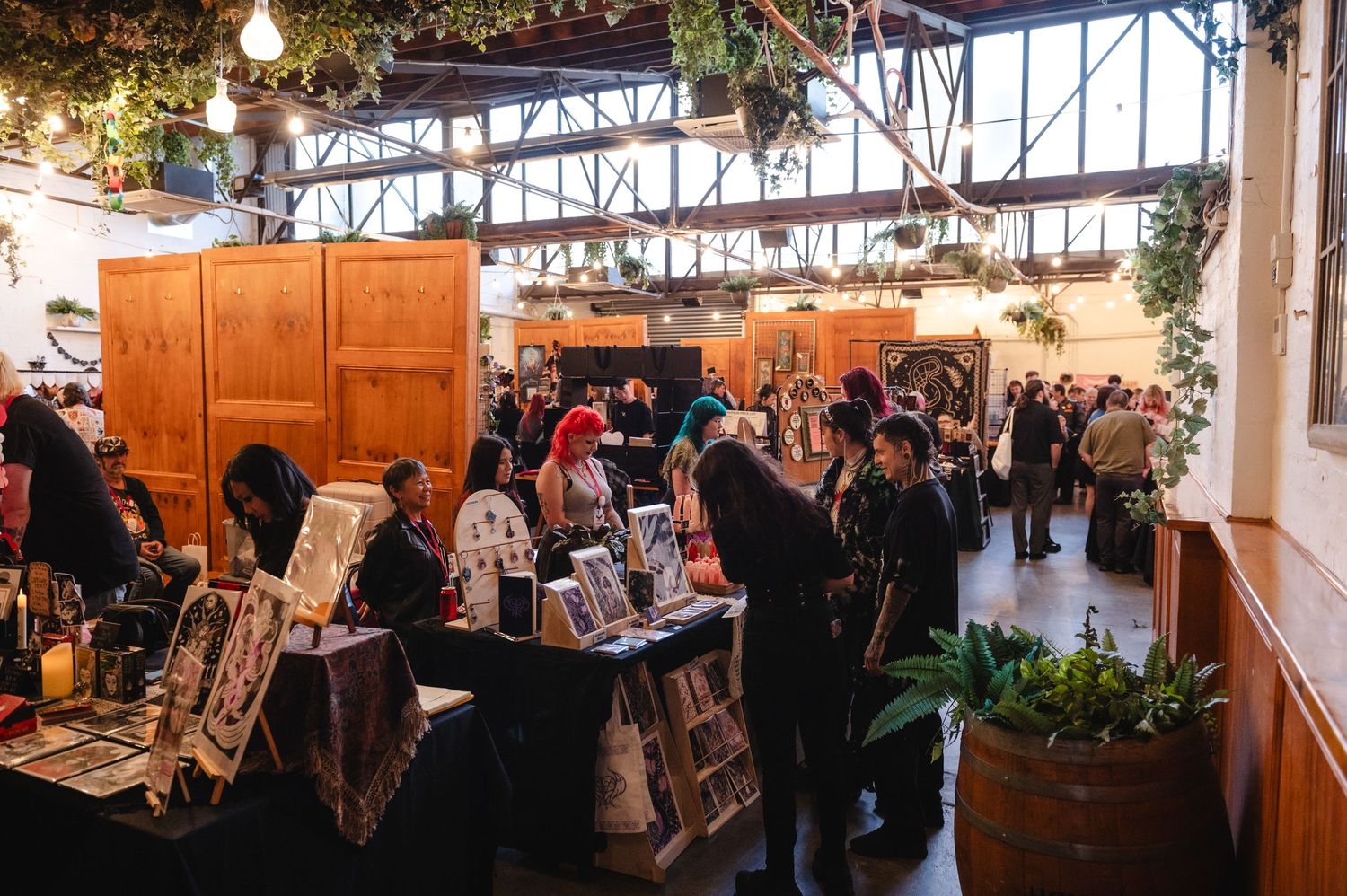 Busy indoor artisan market with vendor tables displaying handmade goods, shoppers browsing, and lush hanging plants decorating the venue.