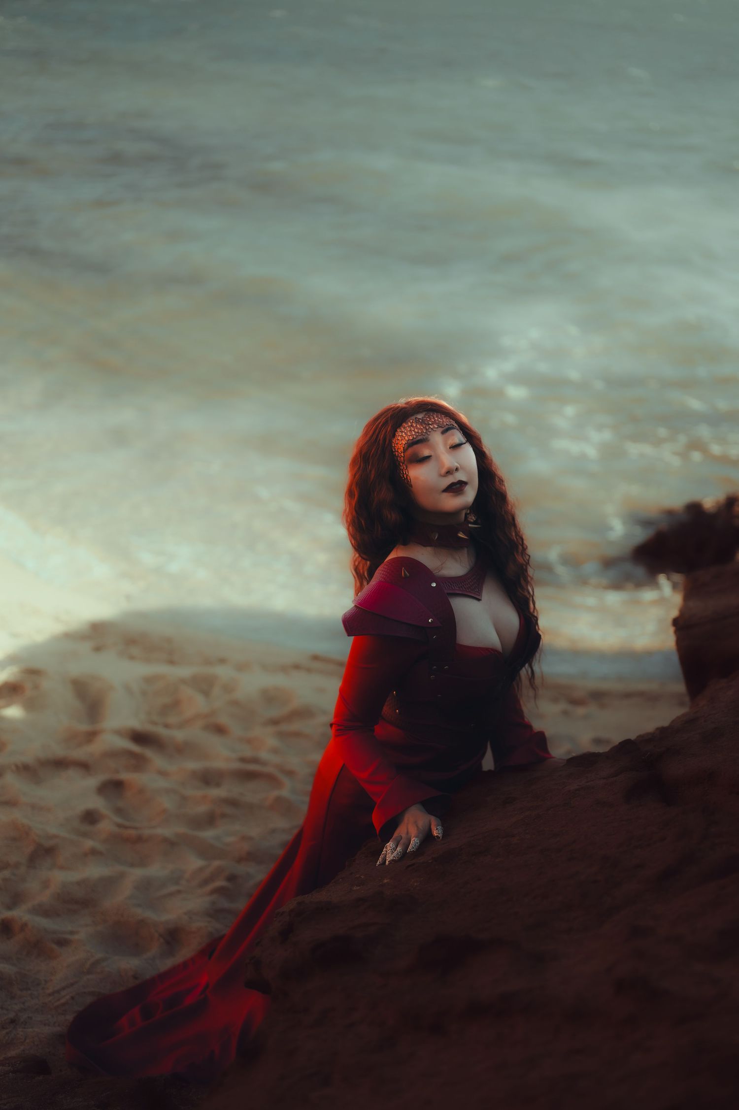 Figure in red dress lying dramatically on rocky shore at twilight.