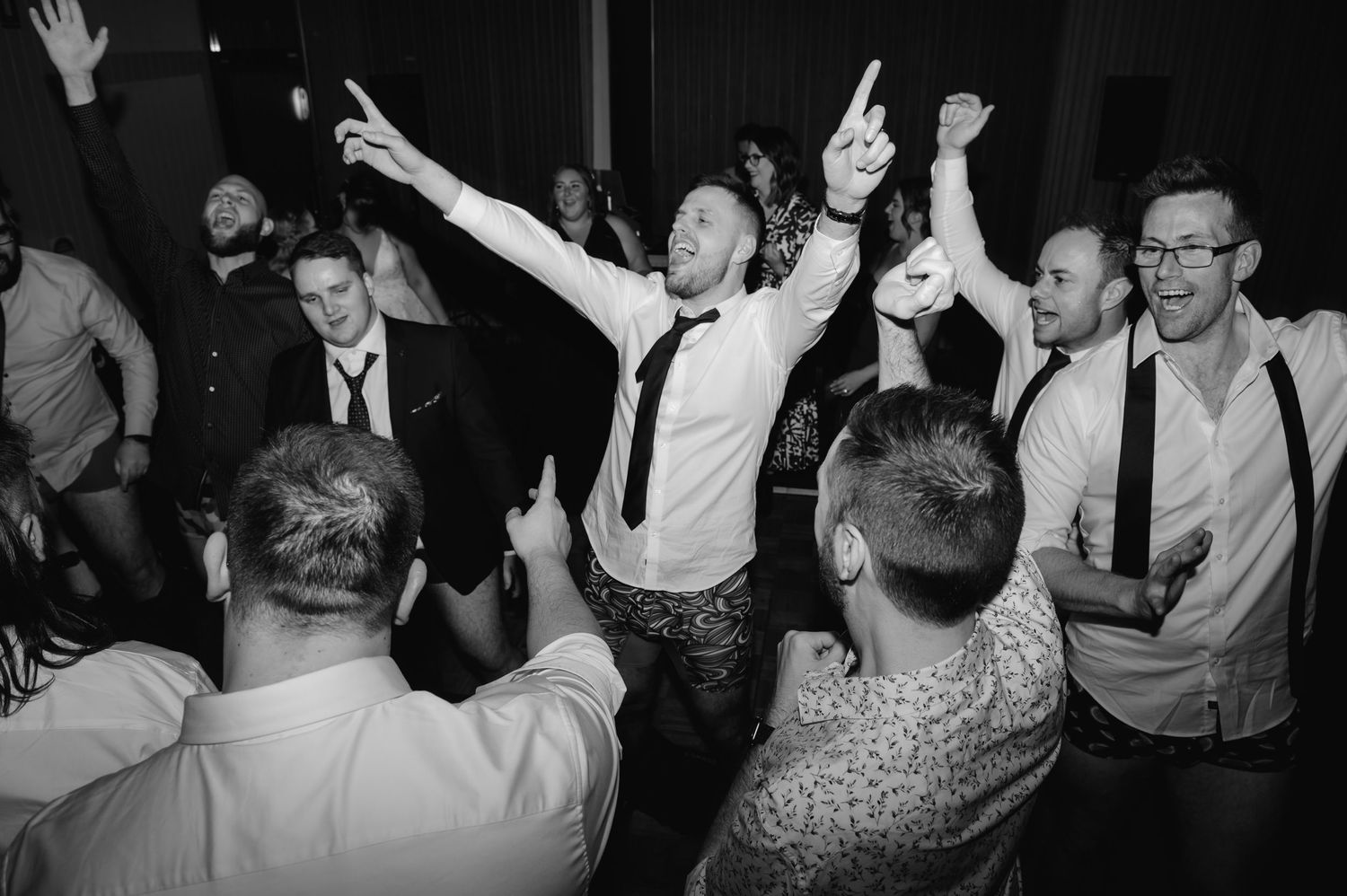 Enthusiastic wedding guests with raised arms dancing together at a reception celebration.