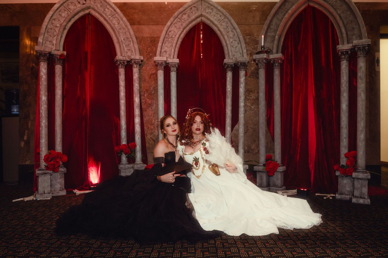 Gothic ballroom scene with elegant dancers in period costumes beneath ornate arched architecture and red lighting.