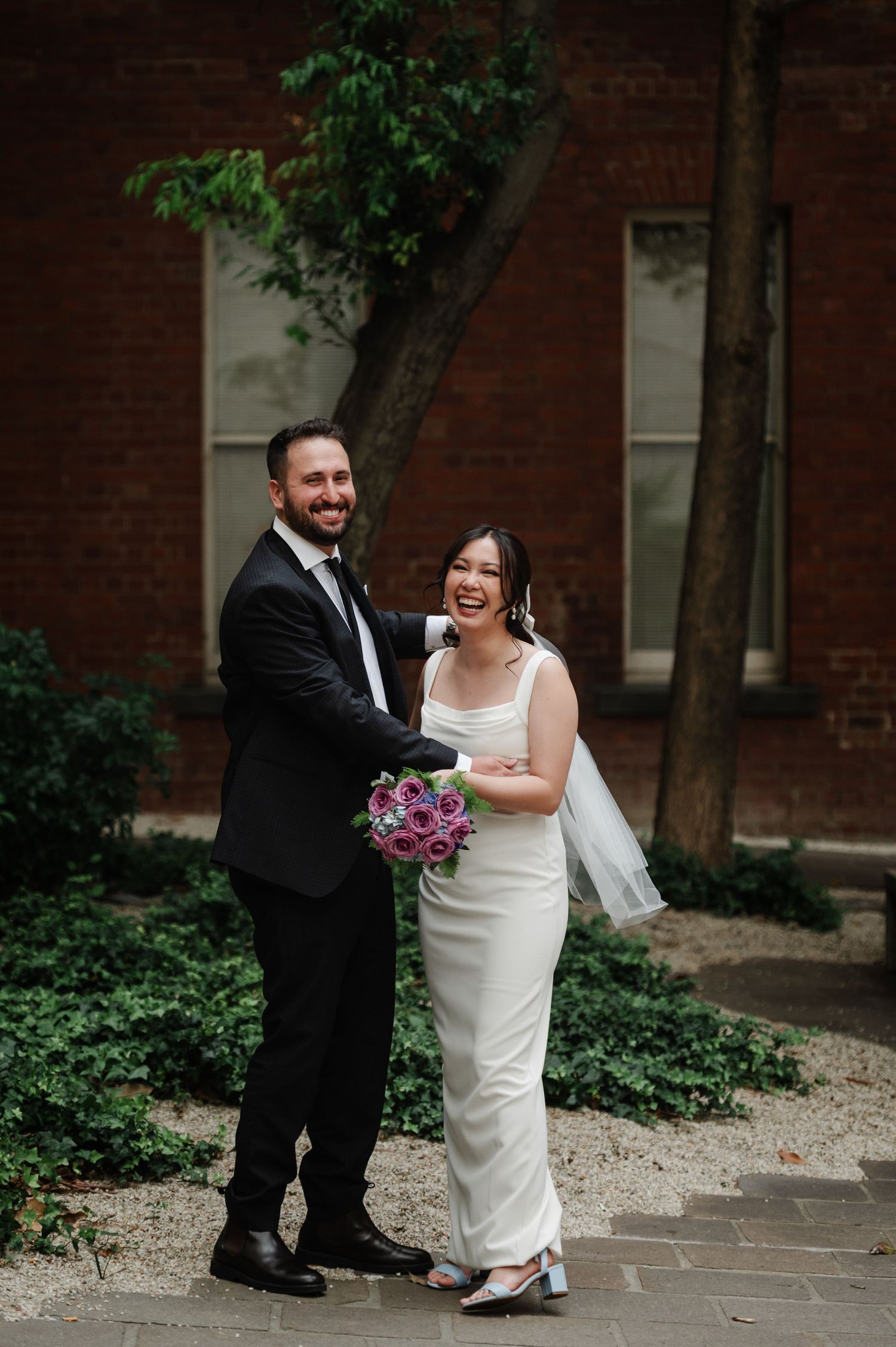 Wedding couple laughs together during outdoor portrait session by brick building.