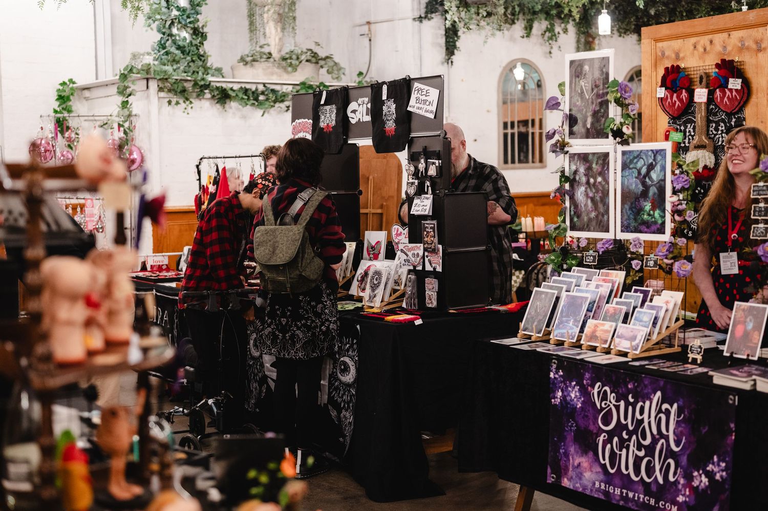 Indoor market booth with Night Witch branding, displaying gothic art prints, handmade crafts, and occult merchandise on draped tables.