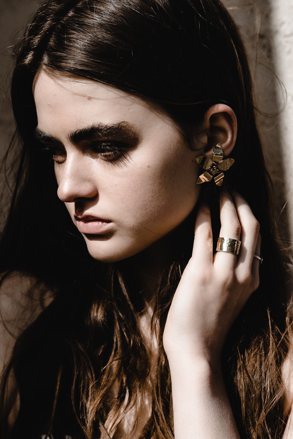 Young woman with dark hair and smoky eye makeup wearing gold geometric earrings and ring, touching her ear.