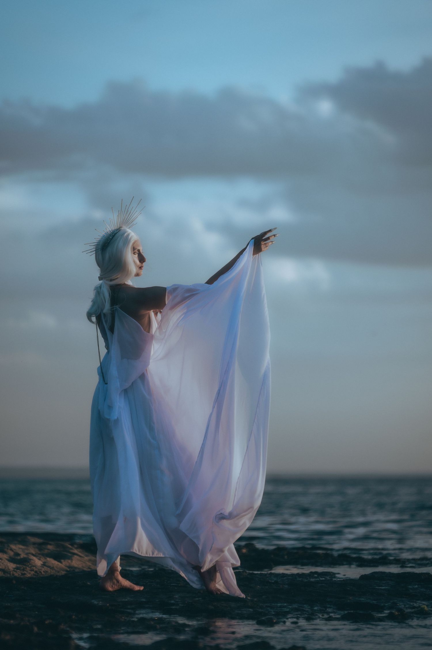 Woman in flowing white sheer dress with silver hair poses dramatically on rocky shoreline at dusk.