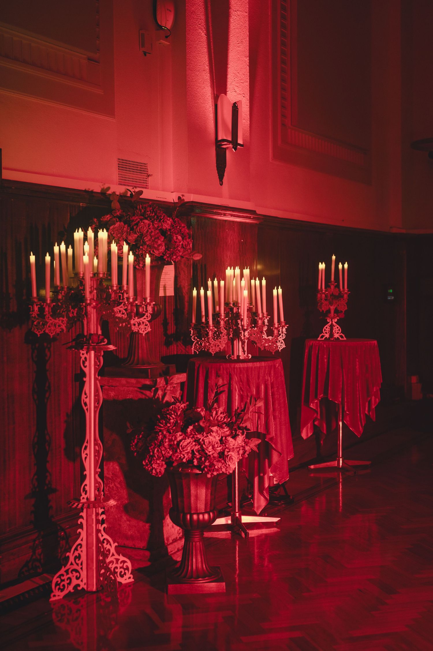 Elegant candelabras with white candles and red floral arrangements create a dramatic gothic atmosphere in deep red lighting.