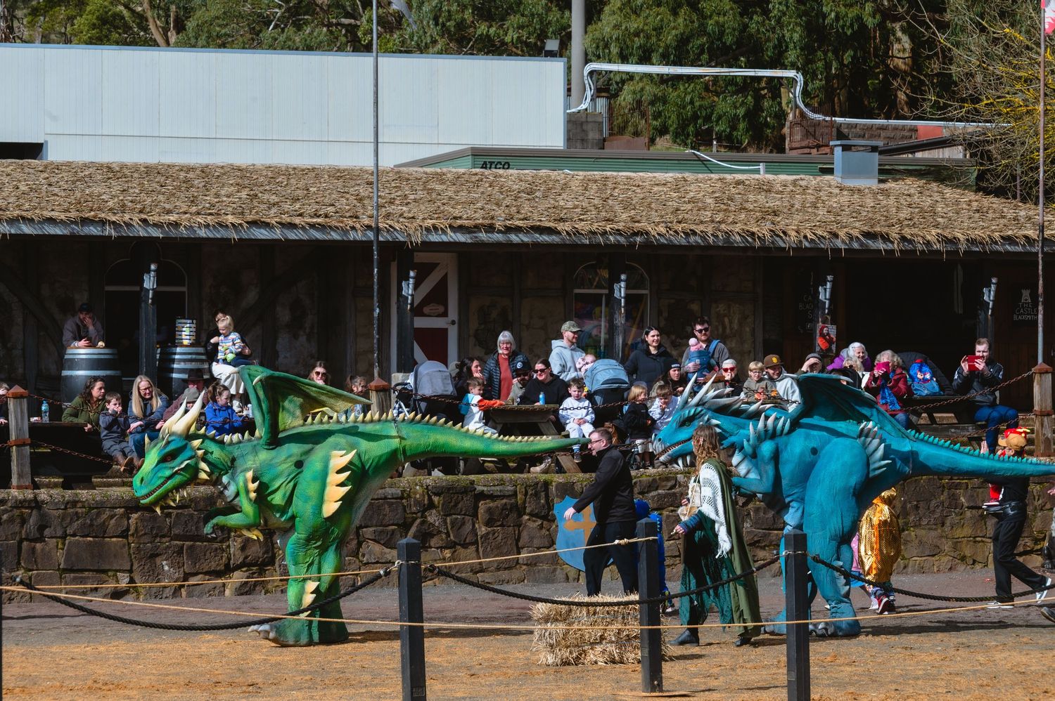 People watch costumed dinosaur characters perform outdoors at a zoo or theme park event on a sunny day.