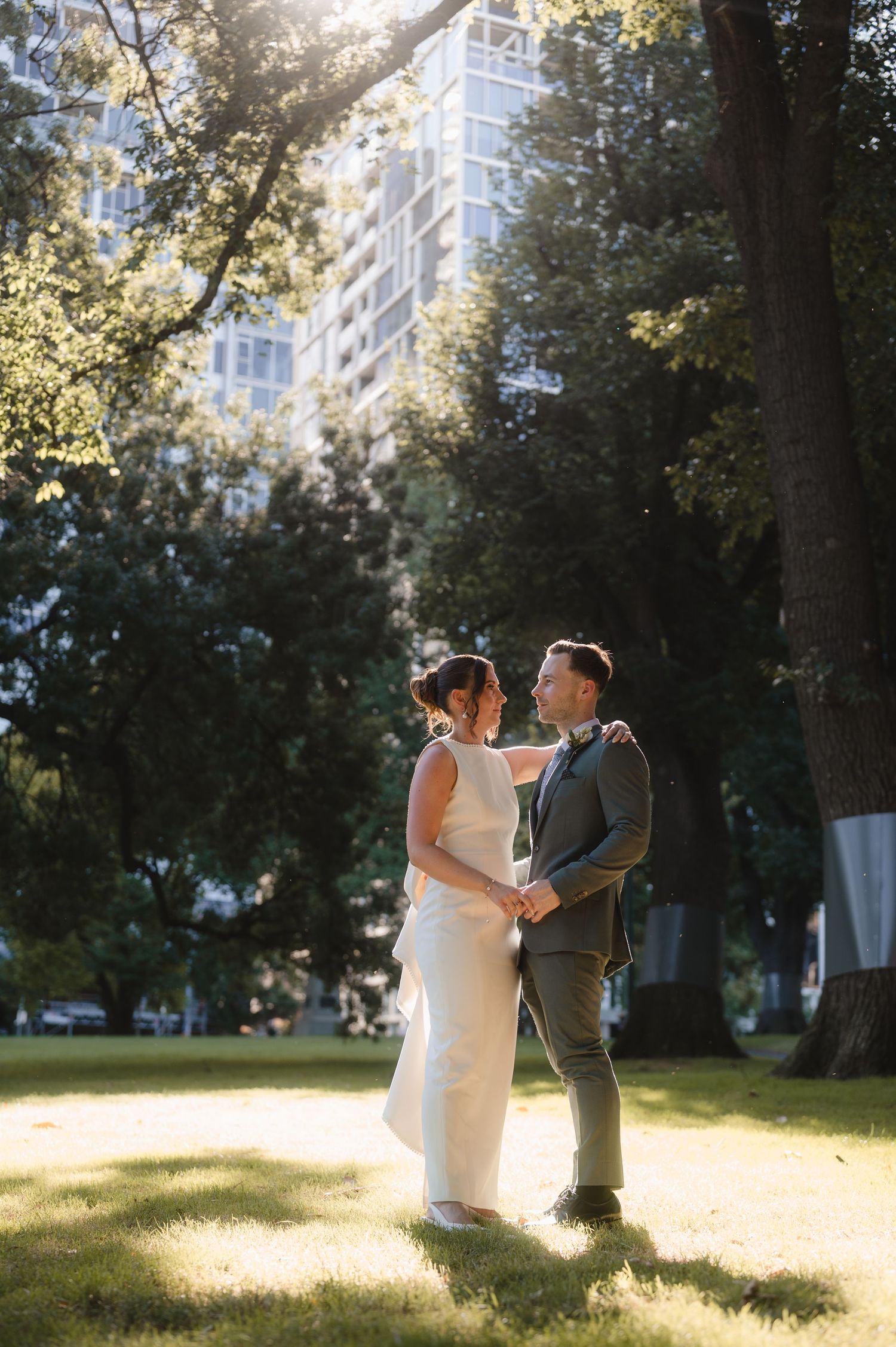 Bride and groom holding hands in sunlit park with trees and city skyscraper backdrop on wedding day.