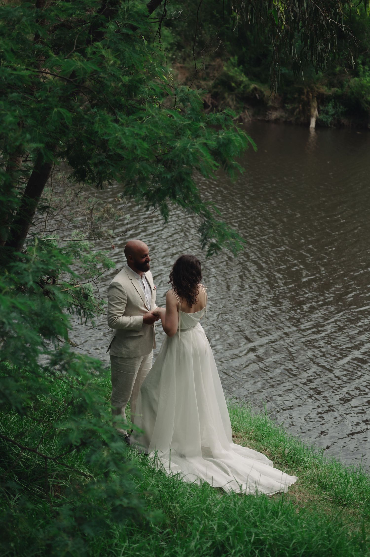 Bride and groom stand hand in hand by a serene river, surrounded by lush green trees on their wedding day.