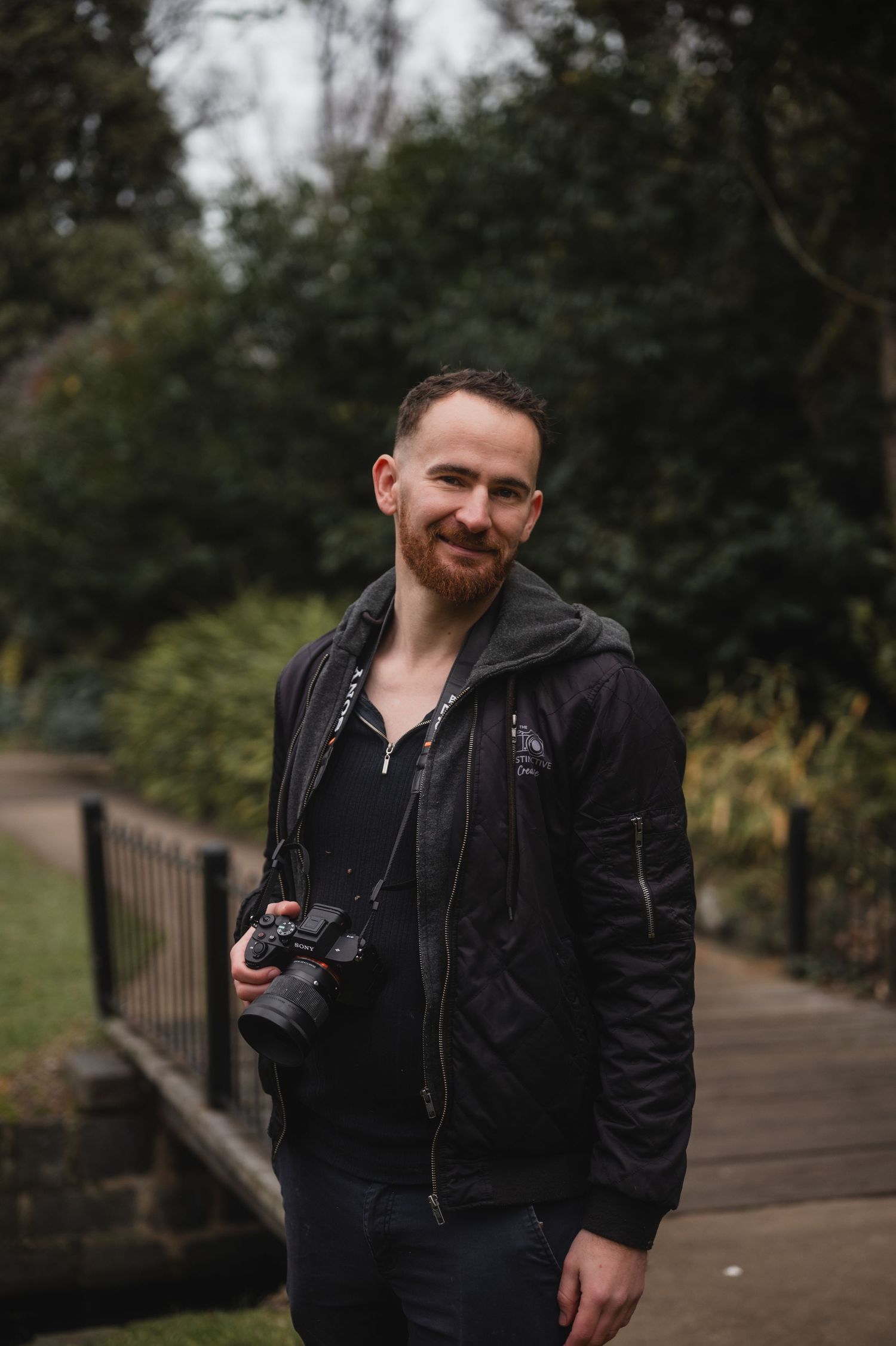 A smiling man with a beard holds a camera in a lush green park, wearing a black jacket.