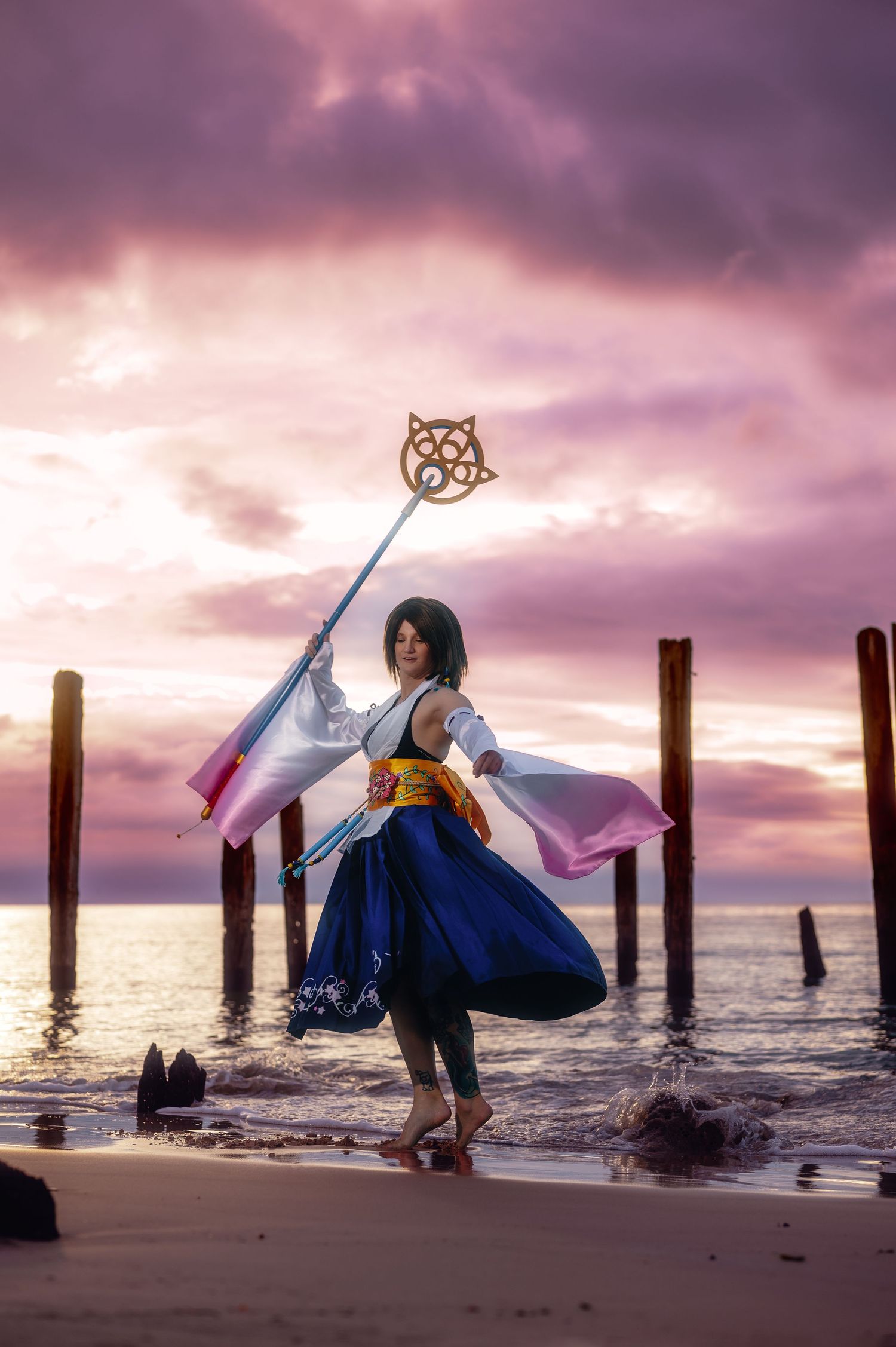 Cosplayer dressed as Yuna from Final Fantasy X holds a staff on a beach at sunset with dramatic purple skies.