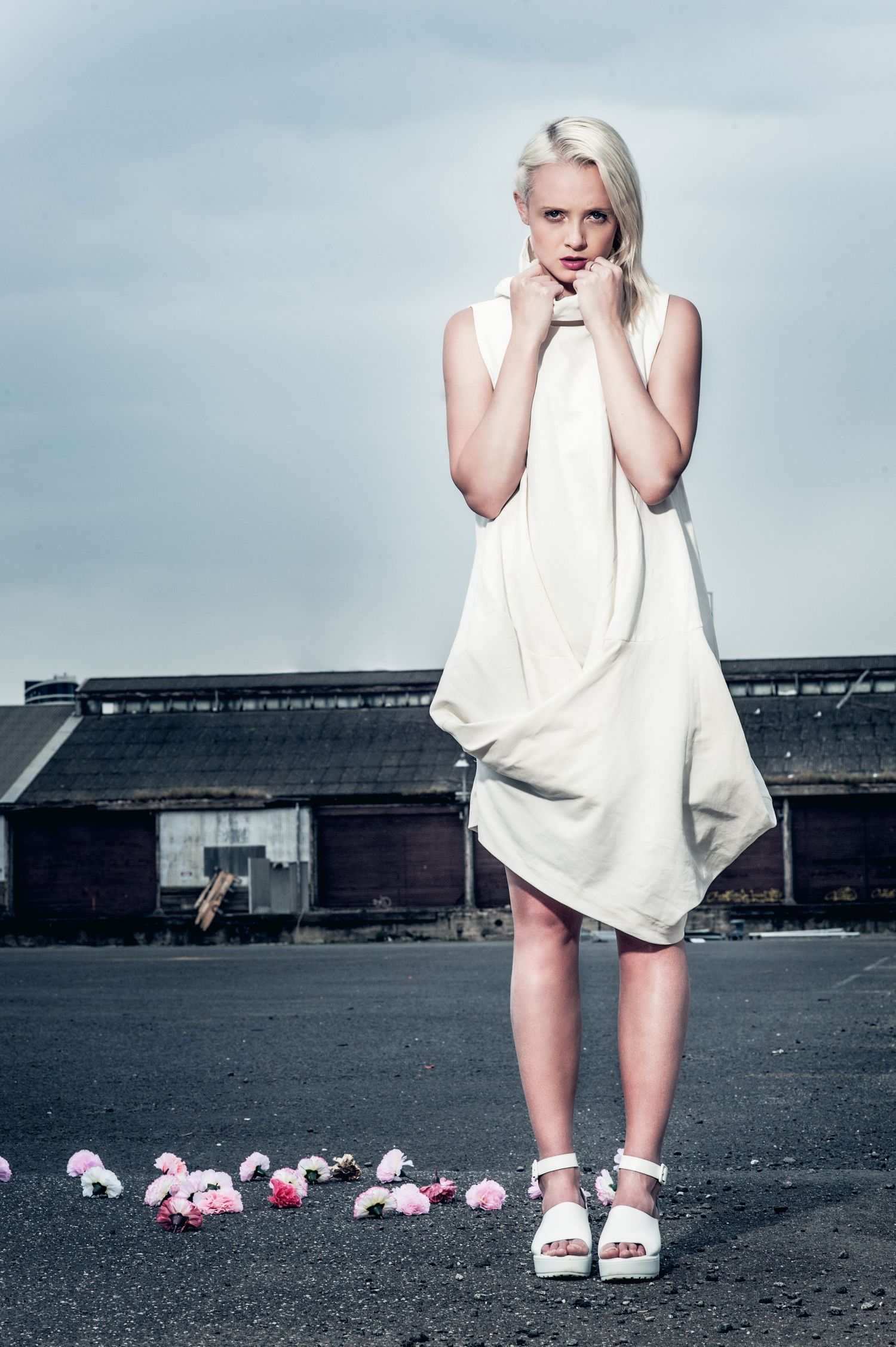 Blonde woman in white asymmetric dress standing on dark pavement with pink flowers, industrial building background.